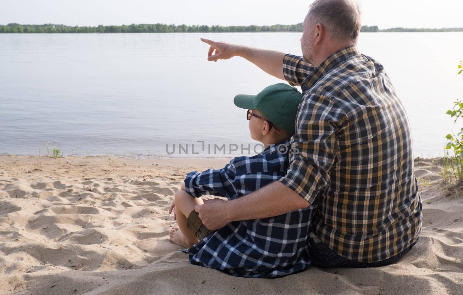 Father's day concept. Rear view of father and son sitting together on the river bank and looking at the water