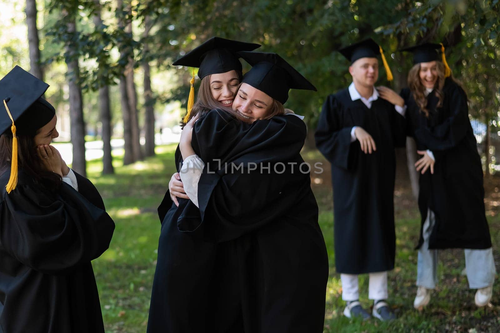 Group of happy students in graduation gowns outdoors. In the foreground, two young girls congratulate each other and embrace