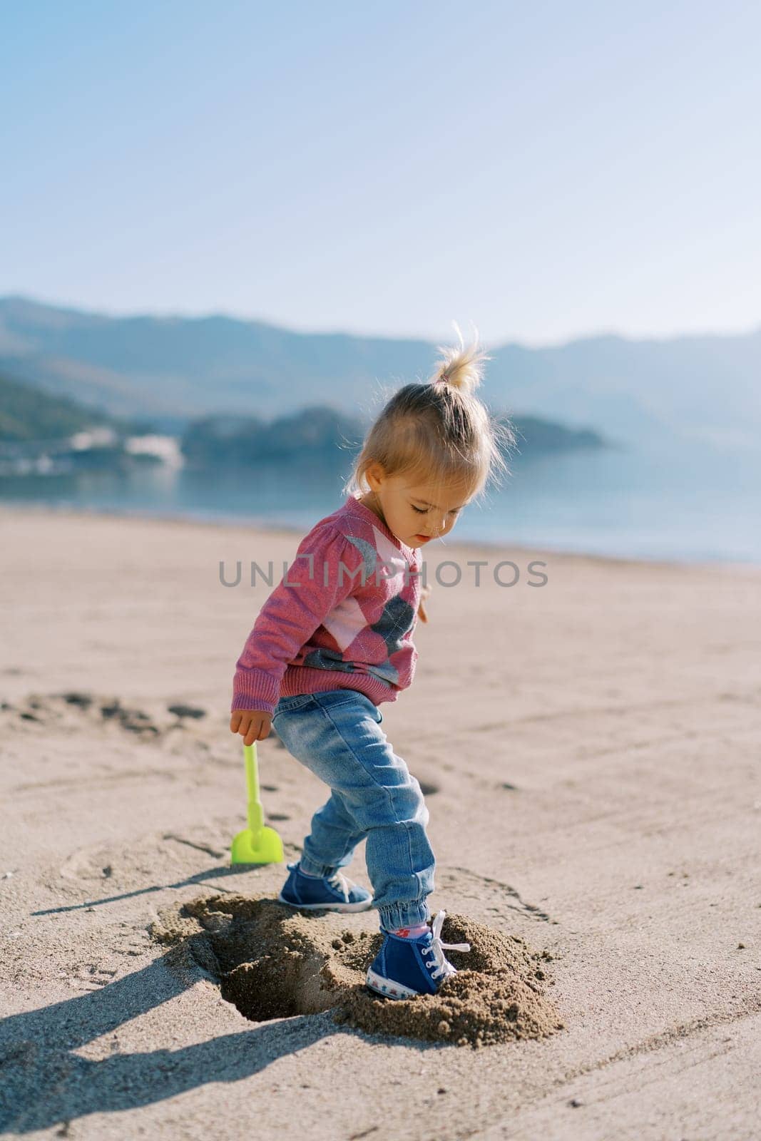Little girl gets her foot into a hole in the sand on the beach near a toy shovel. High quality photo