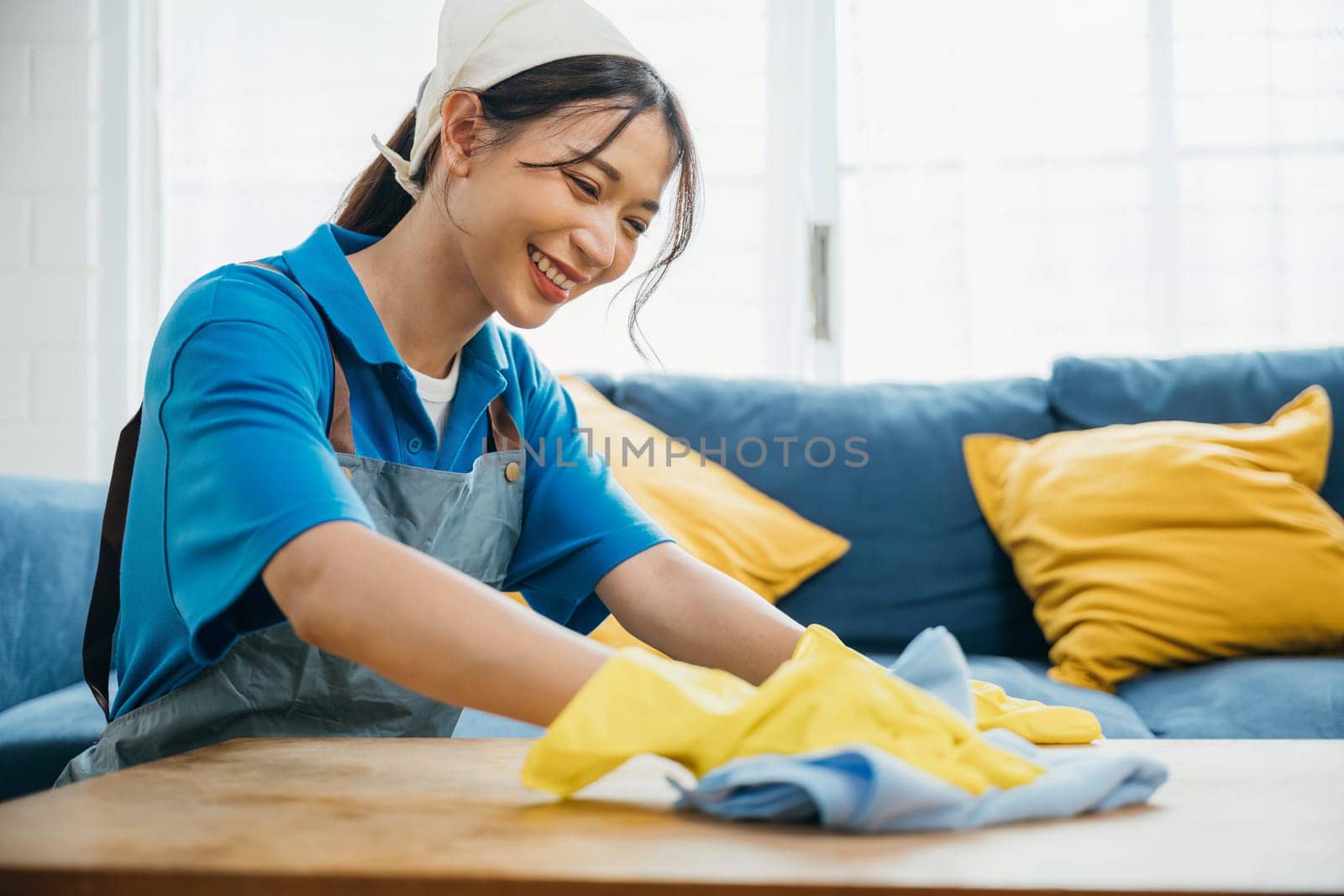 A happy young woman wearing yellow gloves wipes and cleans the table in her living room. Engaged in housework she ensures hygiene and safety showing dedication to home cleaning. maid clean desk. by Sorapop