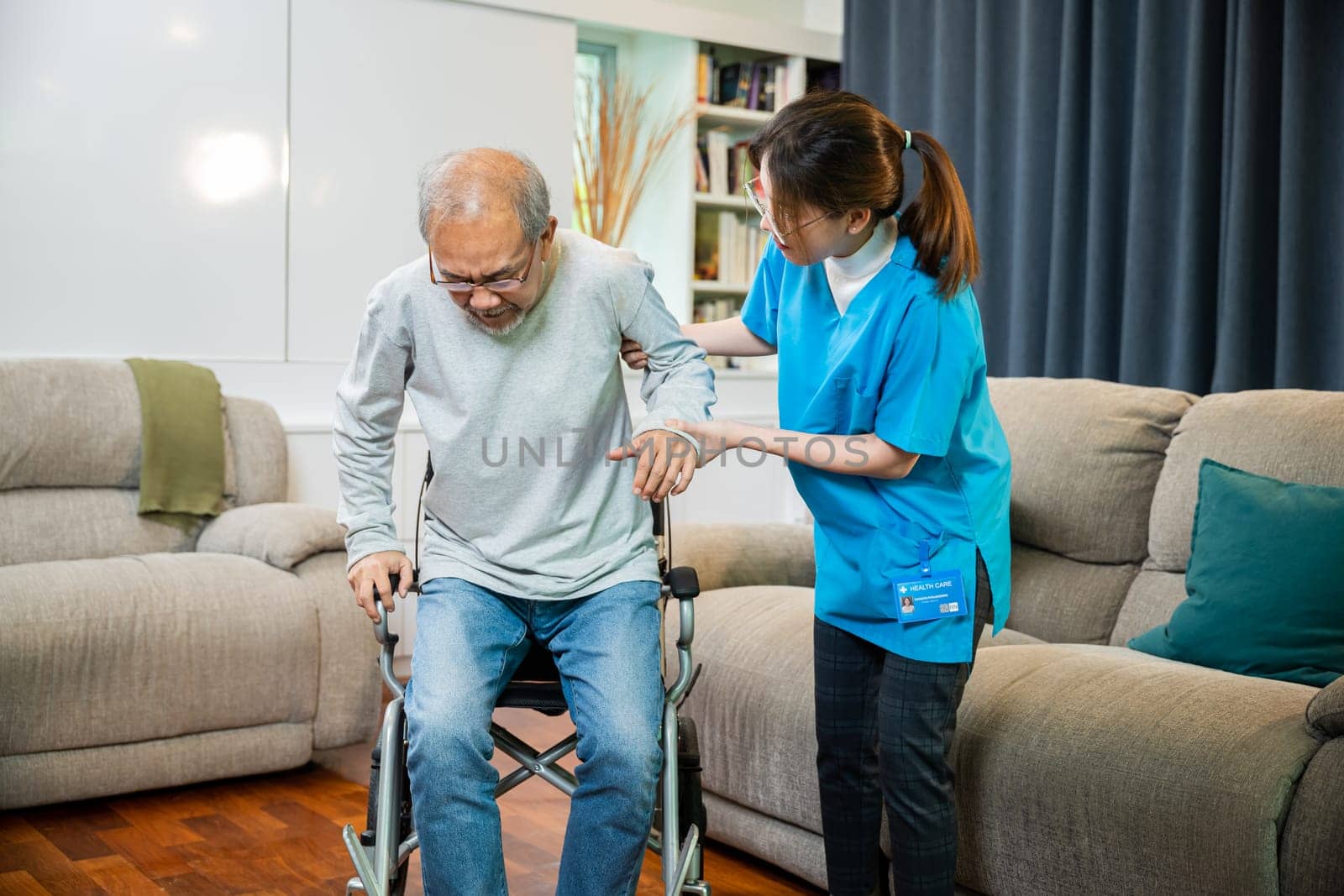 Asian doctor support old man to getting up to exercise, help handicapped elderly stand up, woman nurse assisting helping senior man patient get up from wheelchair for practice walking at home
