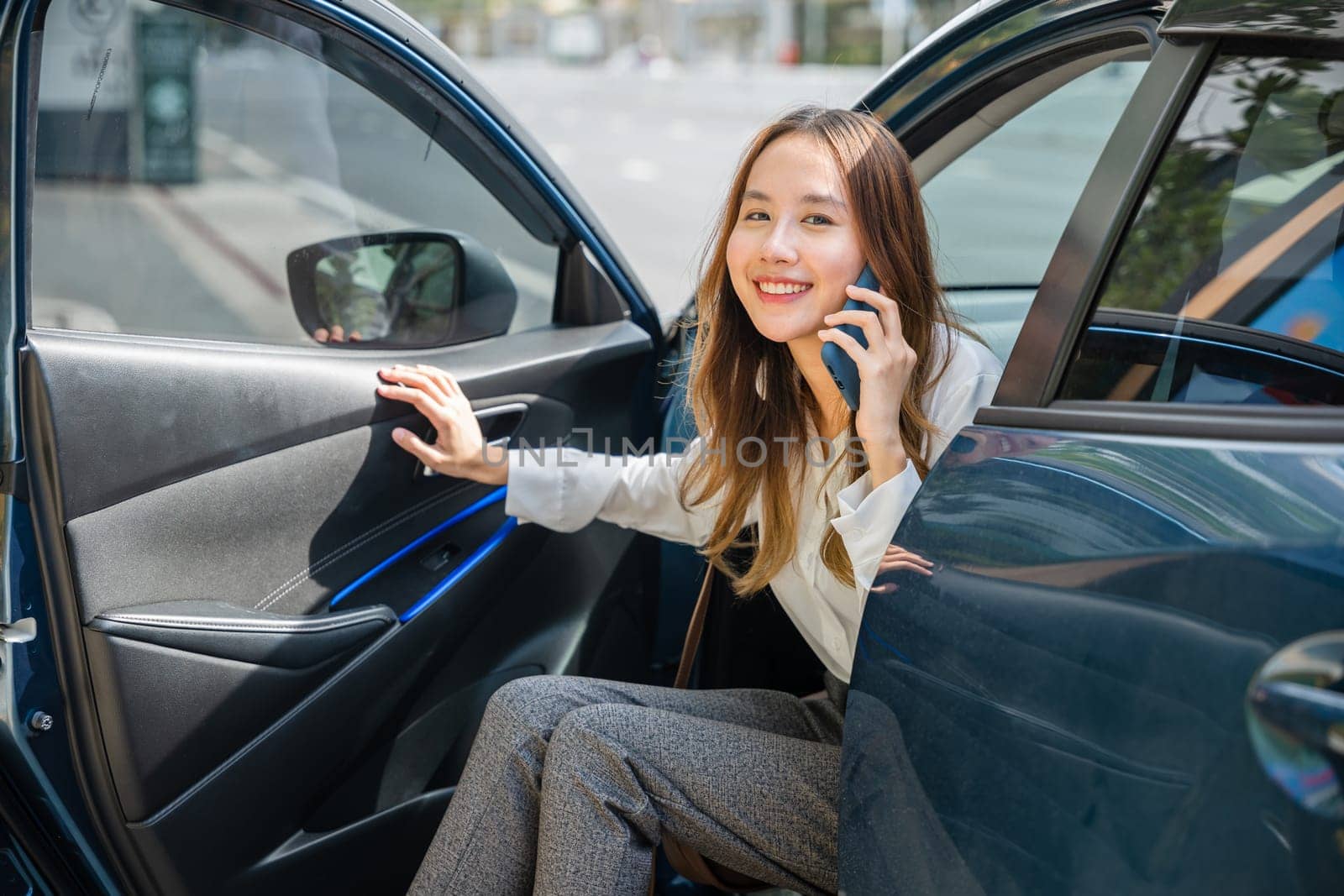 An Asian businesswoman, seated in her luxury car, smiles as she gets a new contract over the phone. Her open car door symbolizes the doorway to business success in the modern city.