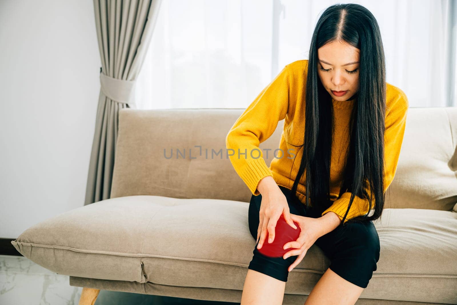 Close-up of a woman on sofa holding her painful knee indicating chronic tendon arthritis. Depicting Health Care and Medical Concept emphasizing tendon inflammation and pain.