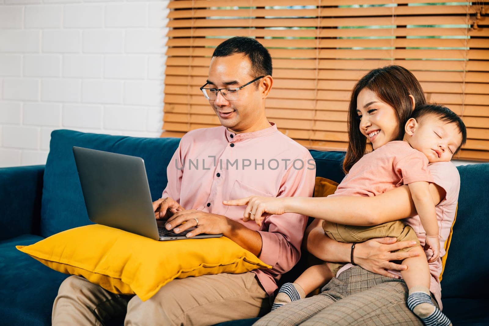 A mother carries her sleeping daughter while her husband works on a laptop depicting the juggling act of raising children and working. Family responsibility and love are evident in this portrait.