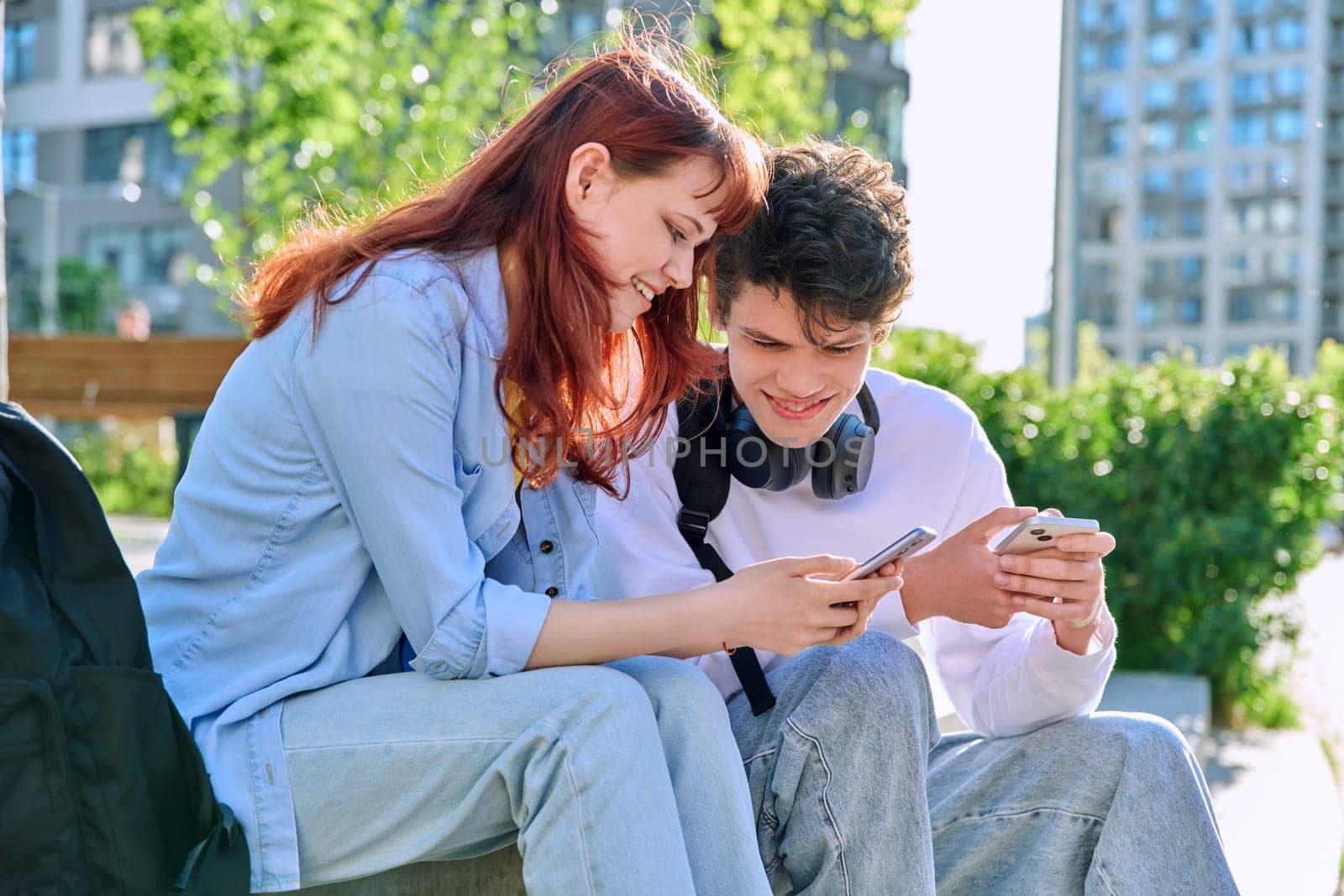 Teenage youth friends guy and girl university college students sitting outdoor on campus steps talking laughing using smartphone. Technology, lifestyle concept
