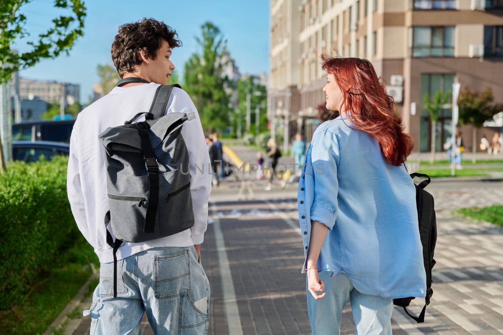 View from back, walking friends, guy and girl university college students with backpacks, talking together along street of modern city. Youth, friendship, communication, urban lifestyle concept