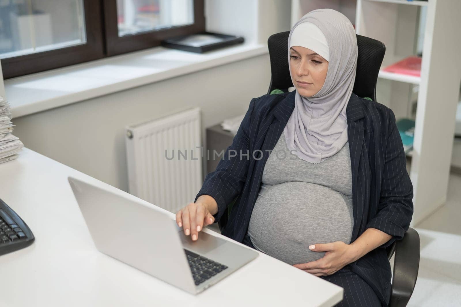 Pregnant Caucasian woman in hijab working at a computer in the office. by mrwed54