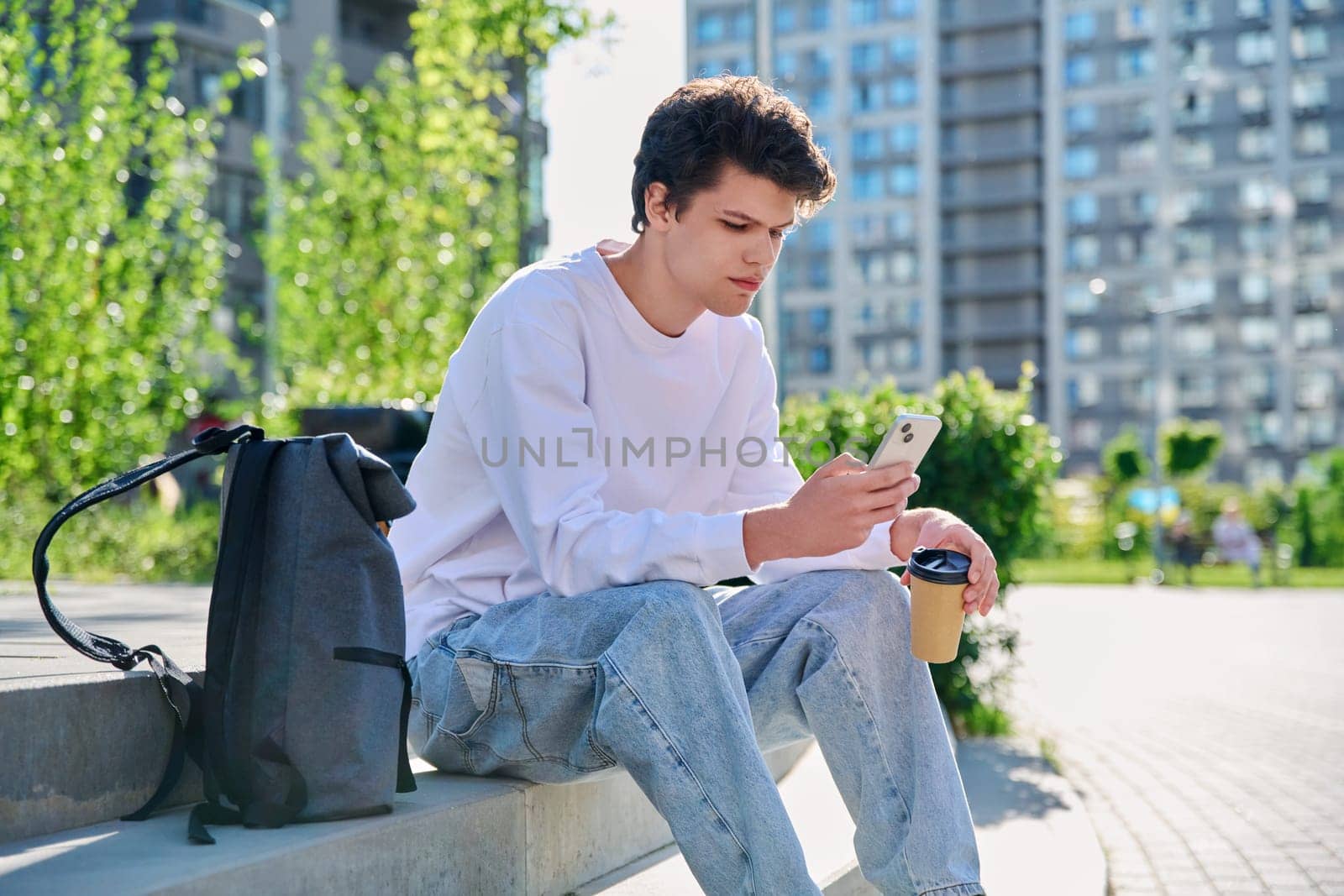 Relaxed resting handsome young male with smartphone, glass of coffee to go, sitting on steps, outdoor. Student 19-20 years old with a backpack, lifestyle, youth concept