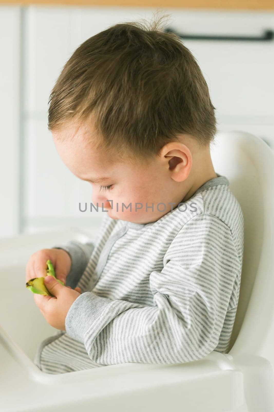Happy baby sitting in high chair eating fruit in kitchen. Healthy nutrition for kids. Bio carrot as first solid food for infant. Children eat vegetables. Little boy biting raw vegetable. by Satura86