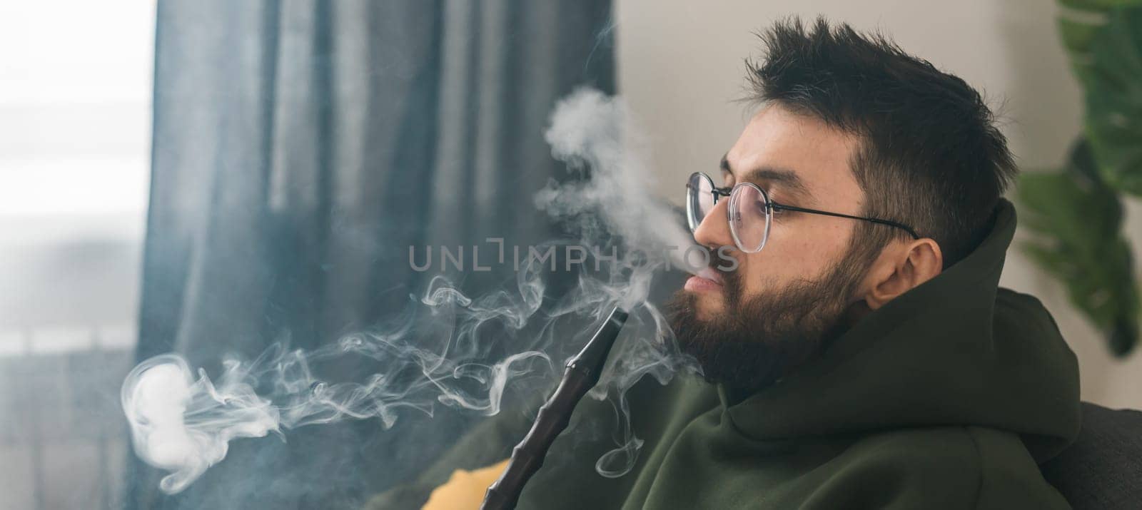 Bearded millennial or gen z man smoking hookah while relaxing on sofa at home - chill time and resting