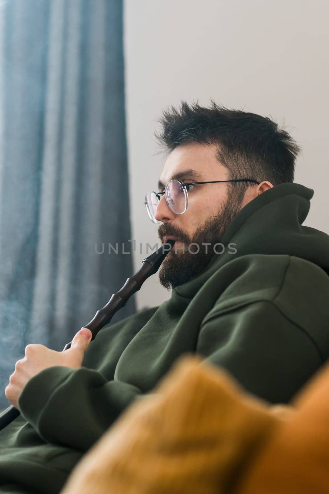 Portrait bearded man is smoking hookah at home and blowing cloud of smoke, copy space and place for text - chill time and resting concept by Satura86