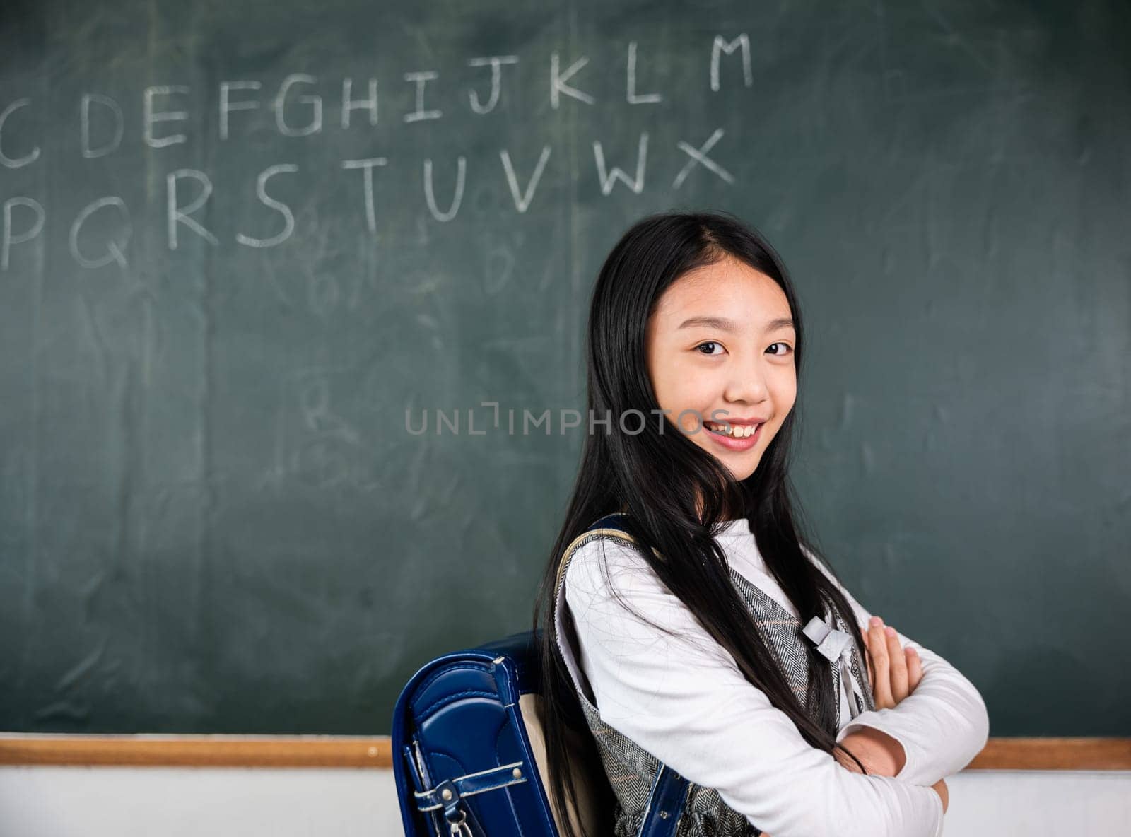 A young girl is smiling and posing in front of a chalkboard with the alphabet on it. Concept of learning and education, as the girl is likely a student in a classroom setting. Back to School