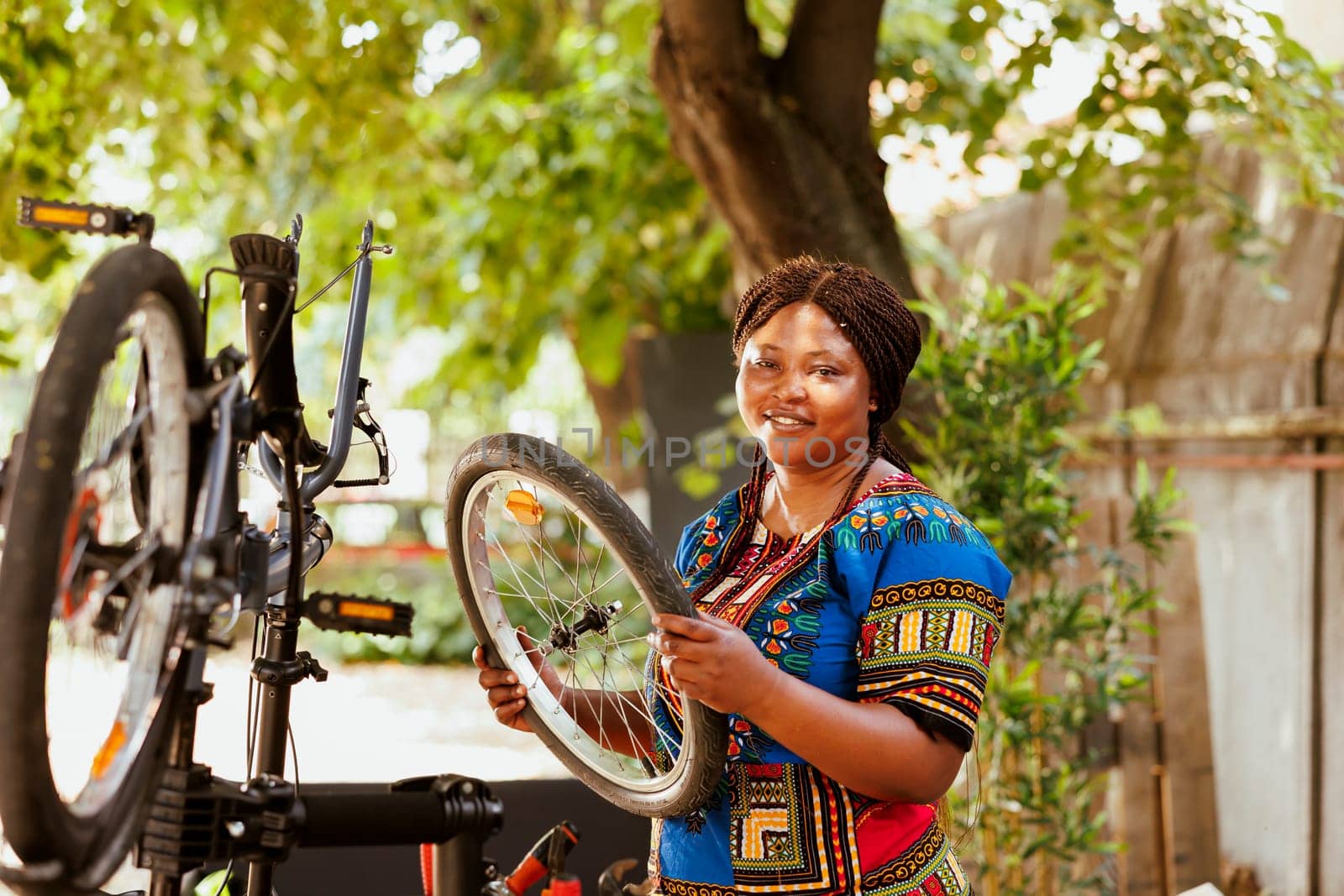 Portrait shot of energetic female cyclist holding dismantled bicycle tire for repair and maintenance in home yard. Active sporty african american woman handling bike wheel for reattachment.