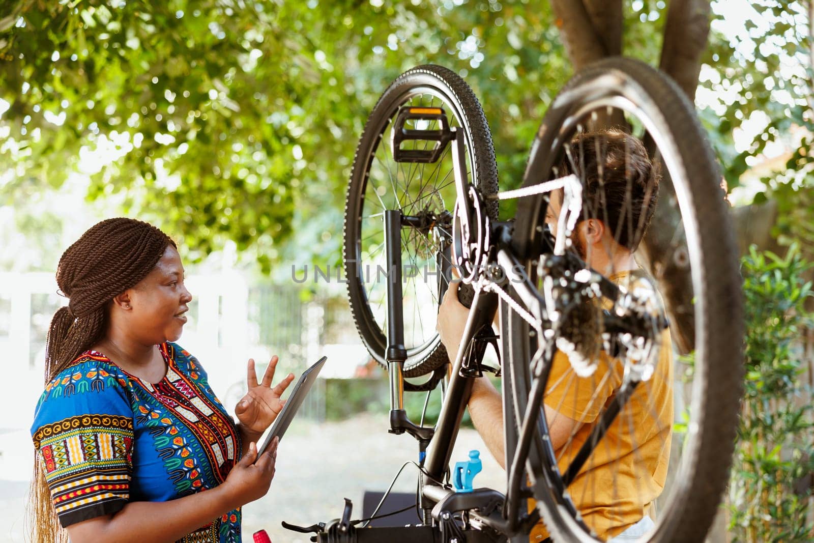 Vibrant black woman happily surfing internet to assist athletic man with bicycle maintenance. Smiling woman with digital tablet gives instructions to active young man to fix bike wheel and chain.