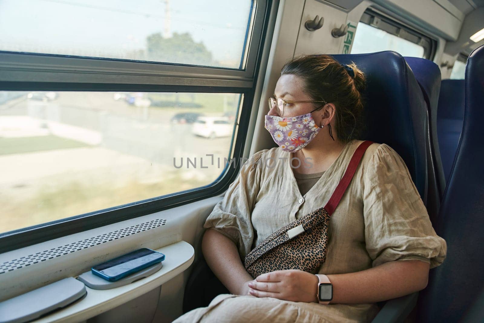 A girl in glasses and a medical mask riding a train and looking out the window by driver-s