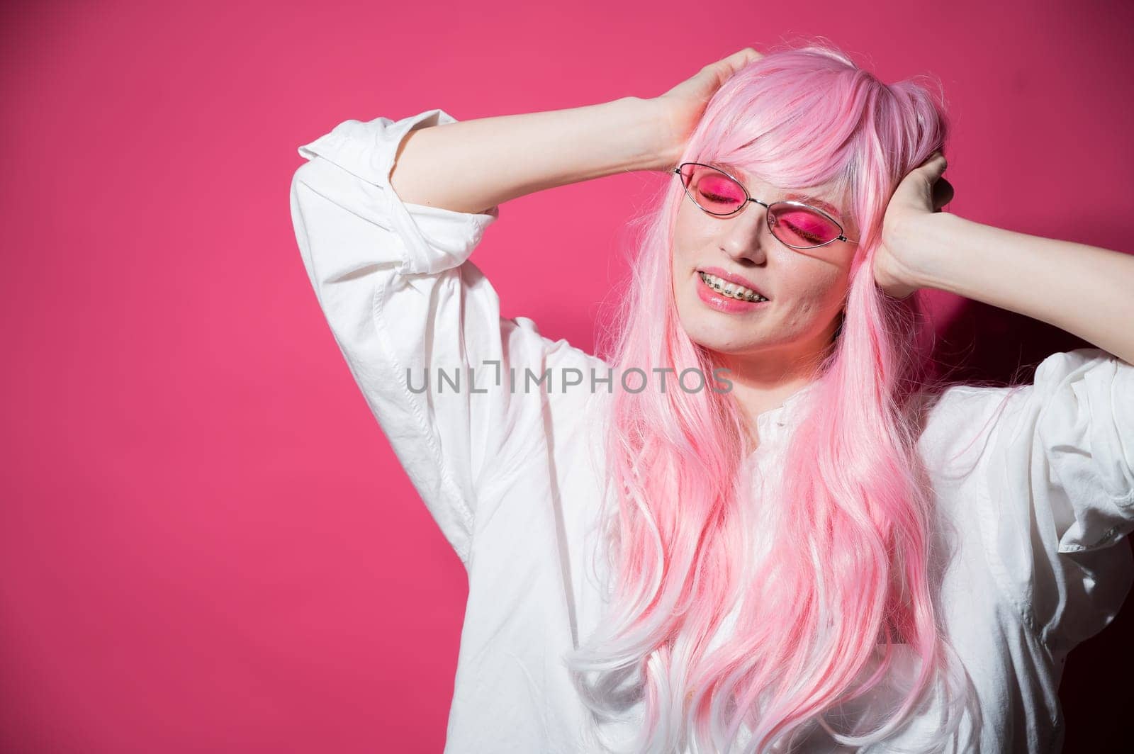 Close-up portrait of a young woman with braces in a pink wig and sunglasses on a pink background. Copy space
