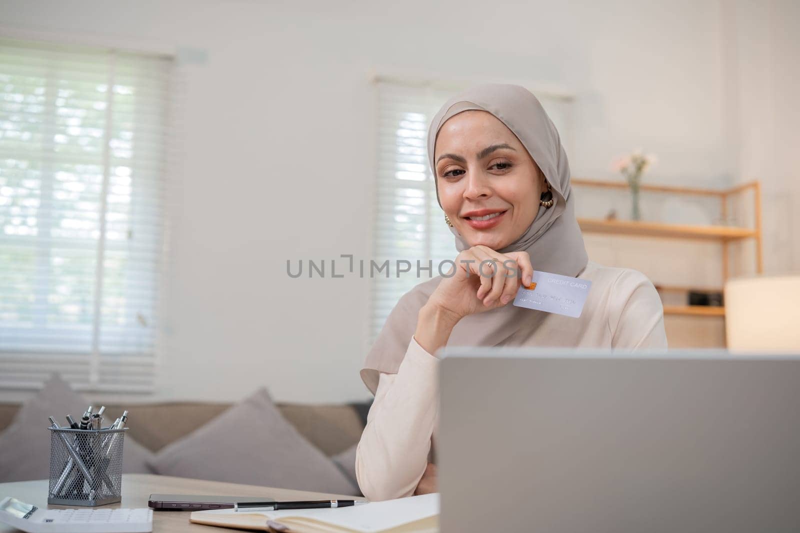 A young Muslim woman wearing a hijab sits contentedly shopping on her laptop, paying through an online banking app and holding a credit card. by wichayada