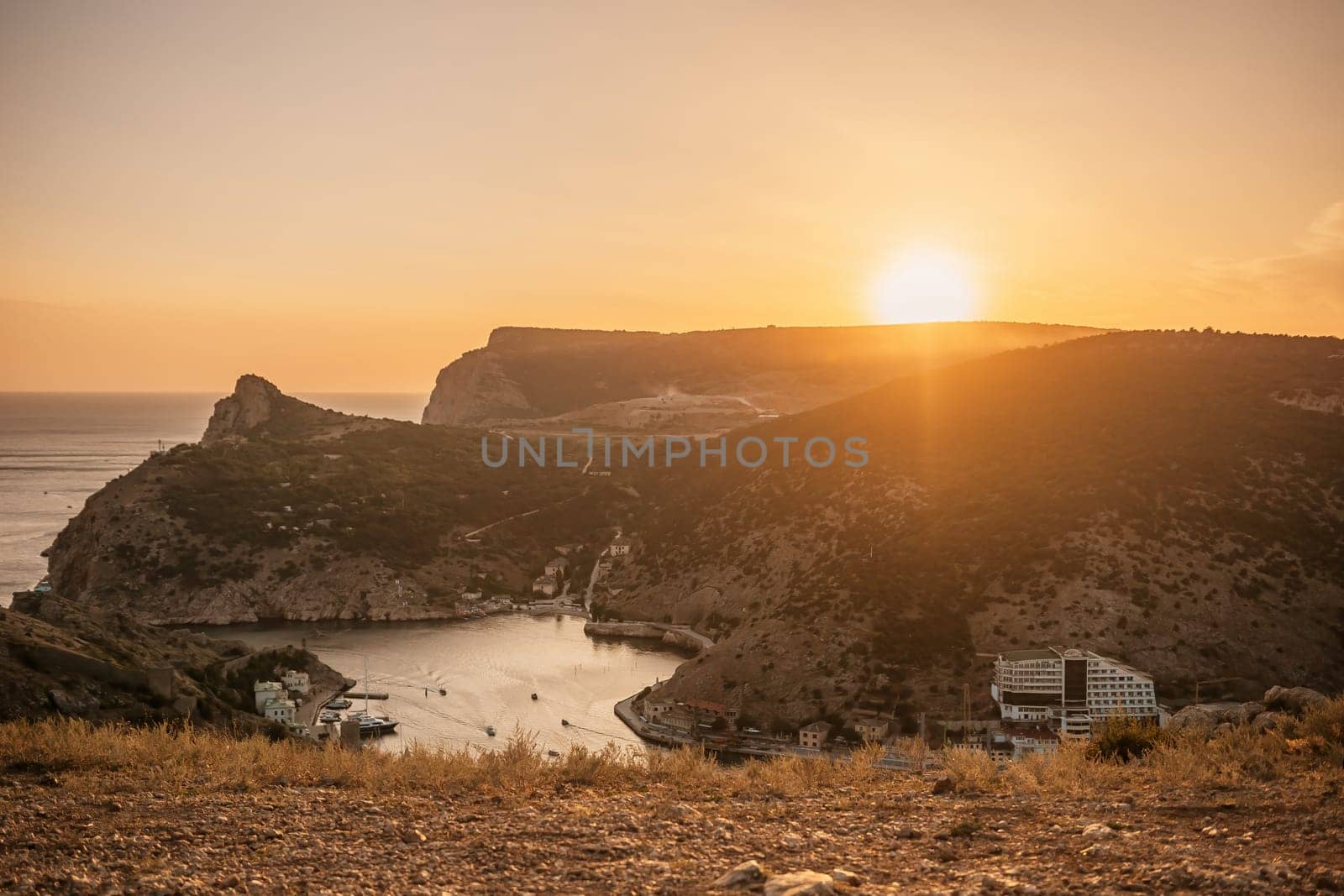 A beautiful sunset over a mountain with a small town in the distance. The sun is setting behind the mountains, casting a warm glow over the landscape. The water in the valley is calm and peaceful