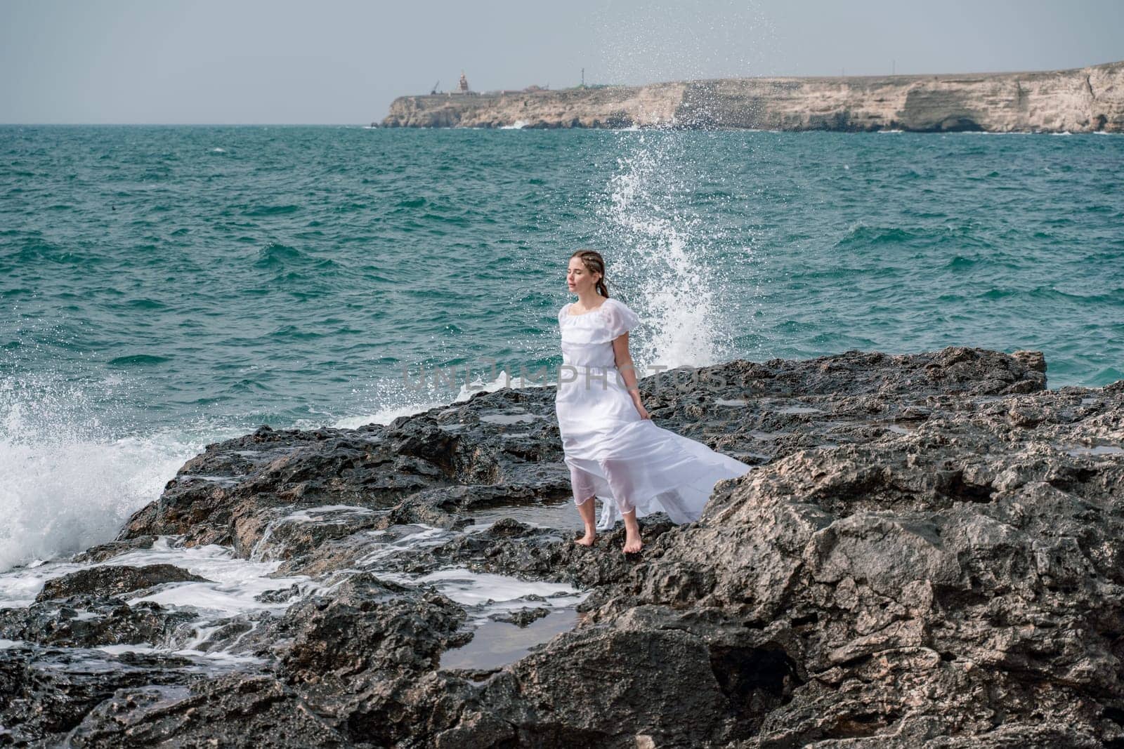 A woman stands on a rock in the sea during a storm. Dressed in a white long dress, the waves break on the rocks and white spray rises. by Matiunina