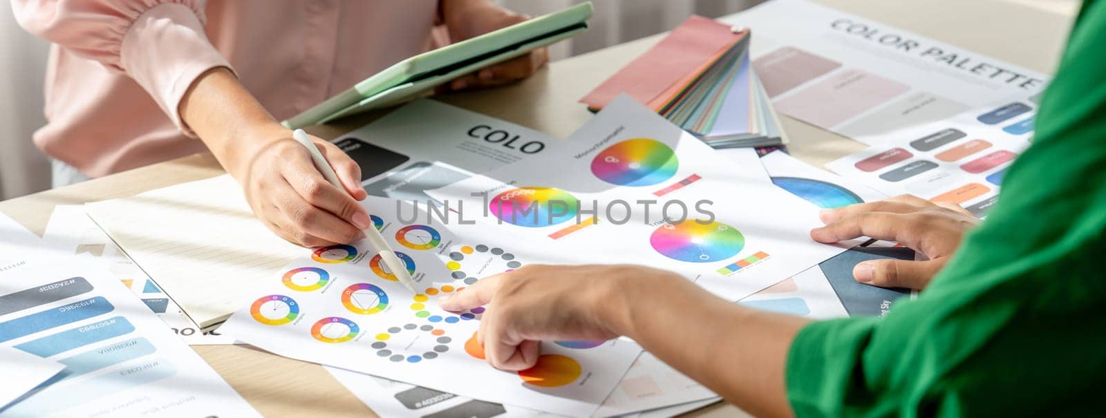 Skilled architect searching data from tablet while selecting an appropriate color from color wheel at table with color palette and document scatter around. Creative design concept. Variegated.