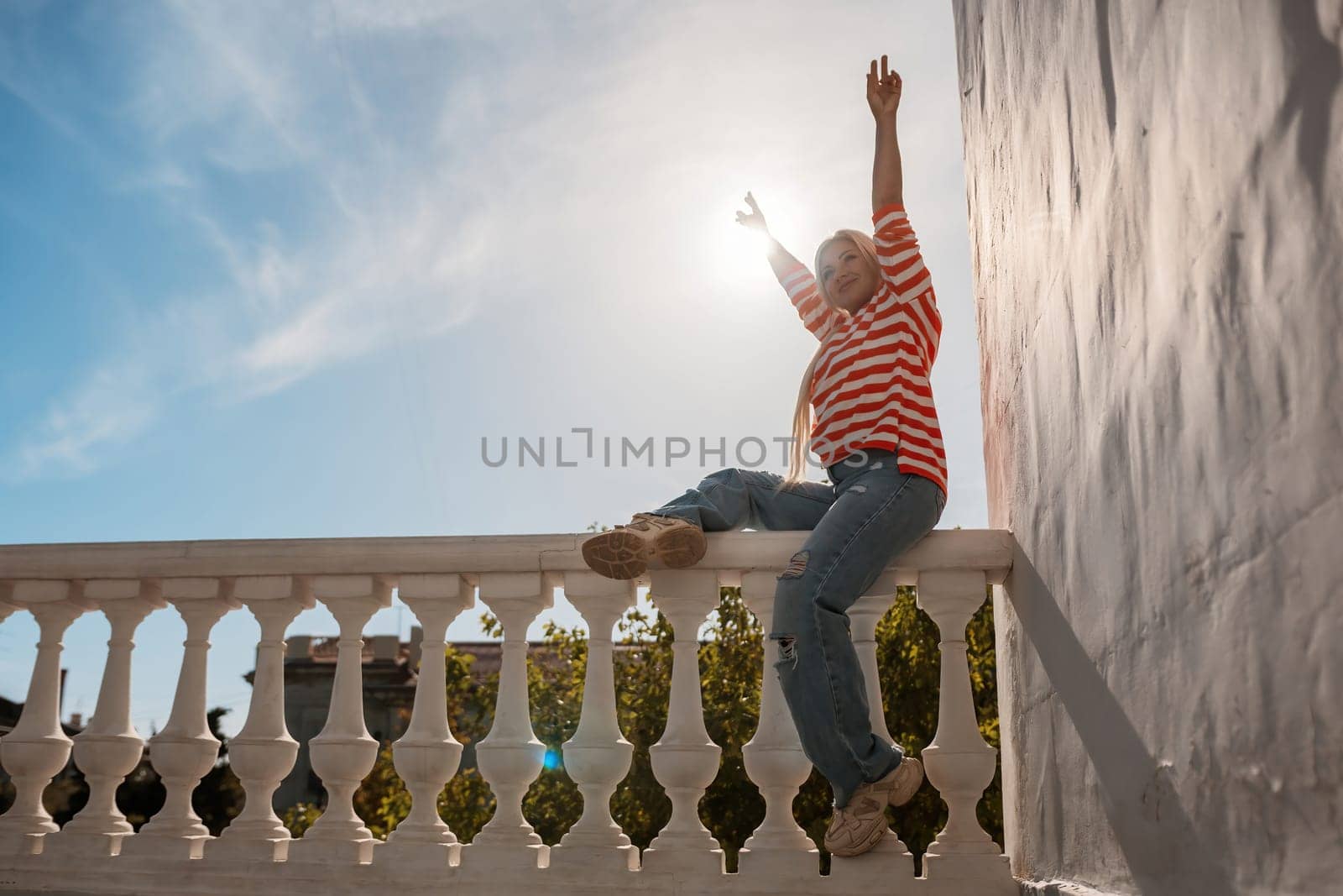 A woman is sitting on a railing with her arms raised in the air. The sun is shining brightly, creating a warm and inviting atmosphere