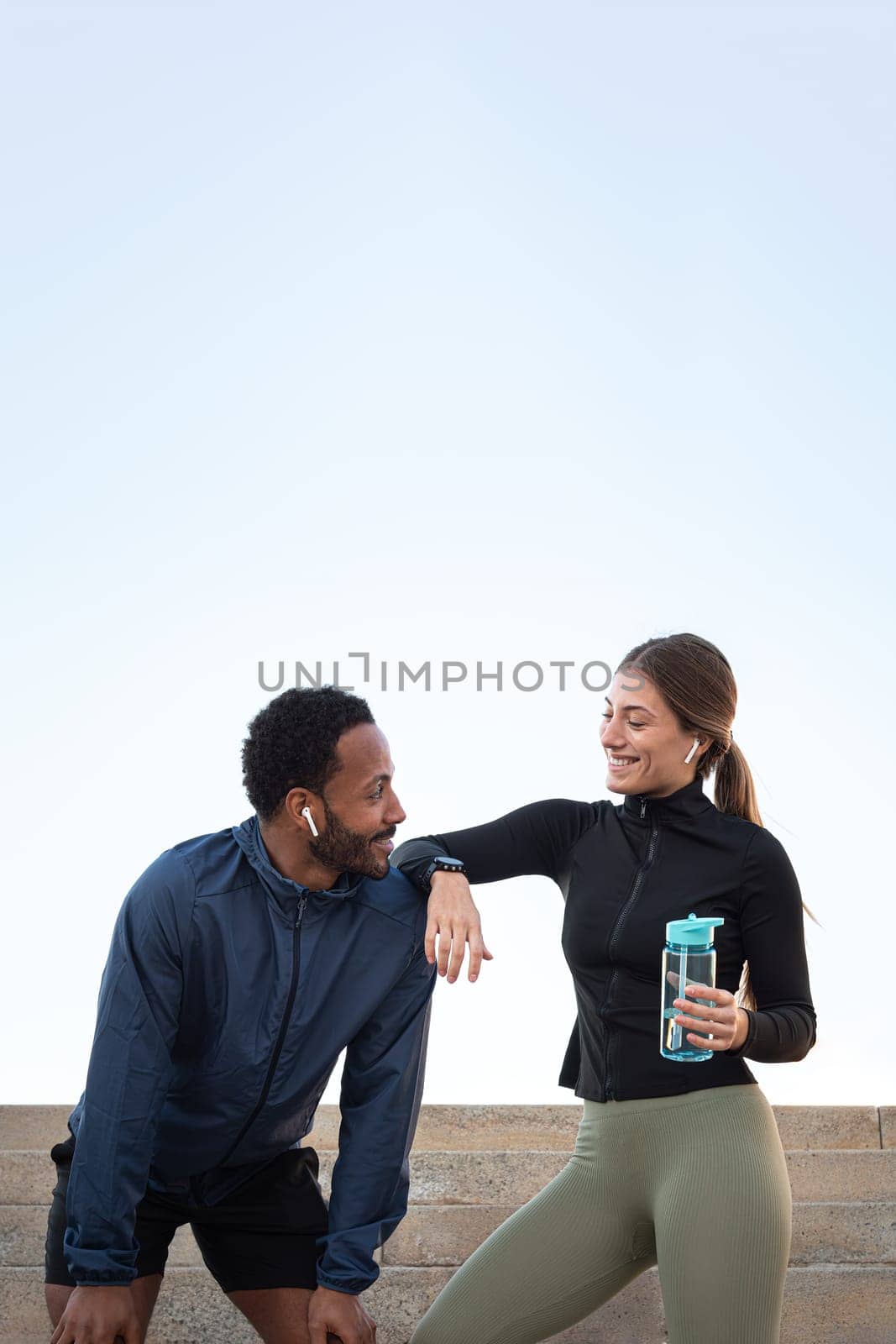 Young happy multiracial couple taking a break after running and working out together outdoors. Female holding water bottle. Fitness and sport concept. Vertical concept.