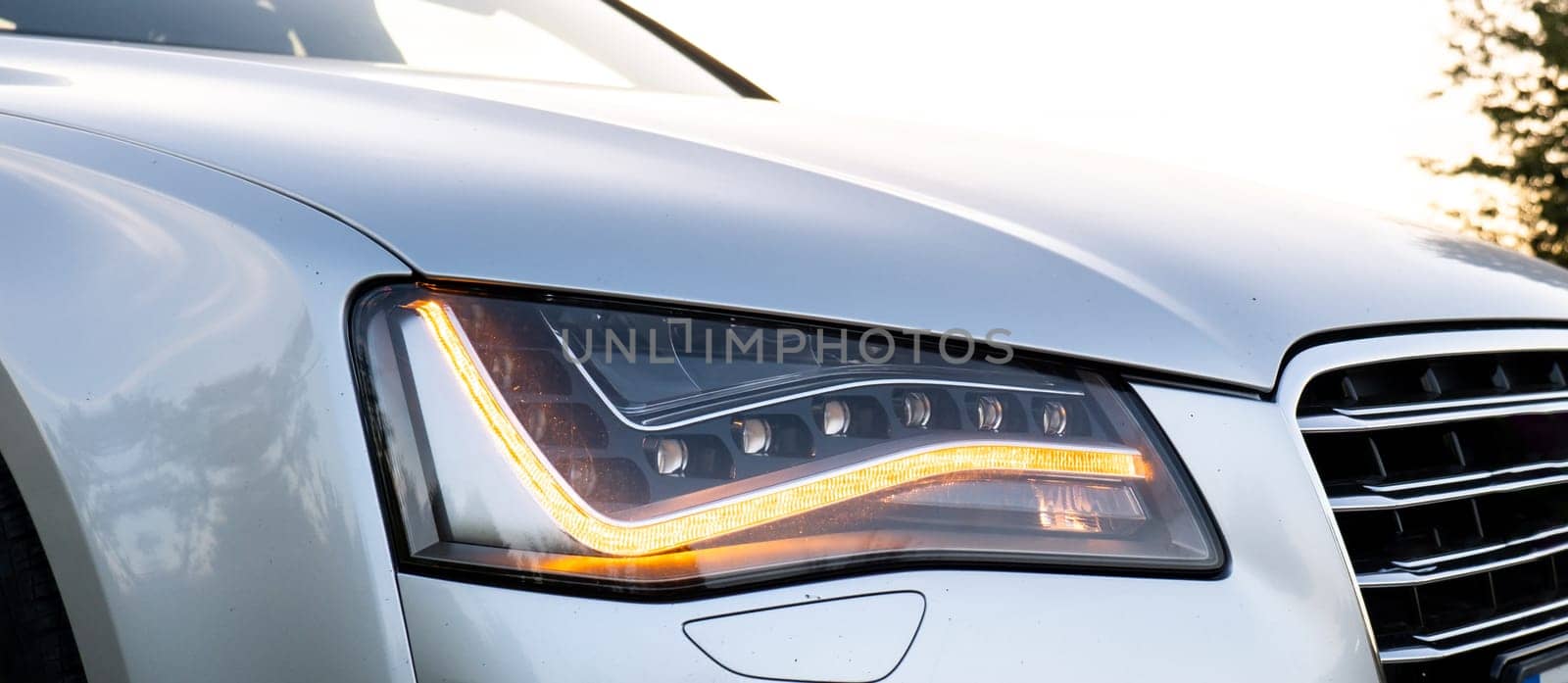 Headlight turned on in Clean car after Washing luxury silver car. Sedan car exterior of modern luxury car during sunset on highway road. Details of front headlamp. Prestige sport by anna_stasiia