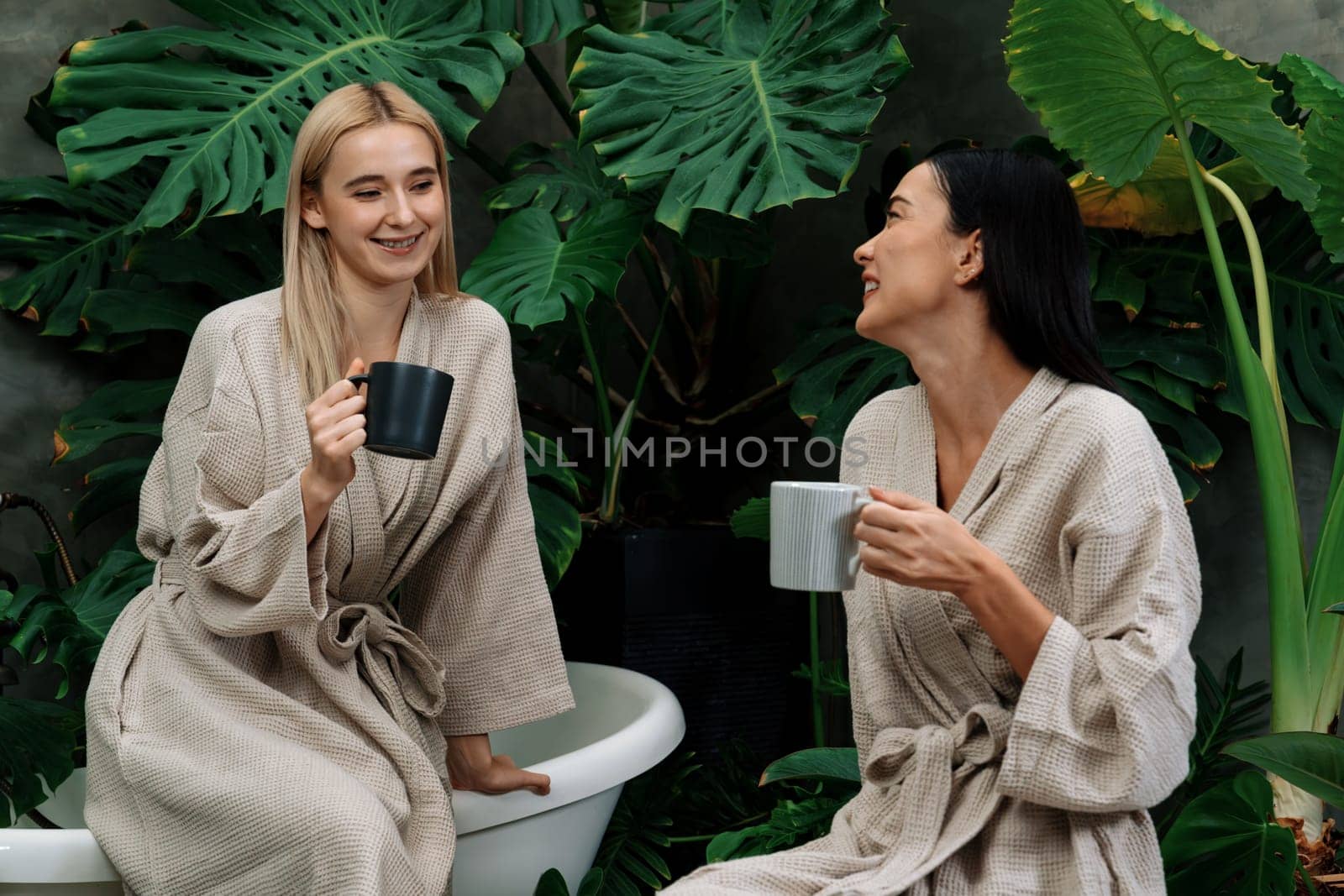 Tropical and exotic spa garden with bathtub in modern hotel or resort with young two women in bathrobe drink coffee, enjoy leisure and wellness lifestyle surround by lush greenery foliage. Blithe