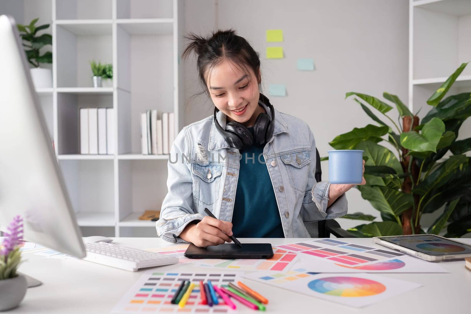 Female designer wearing headphones listening to music while doing graphic design work Choose colors for working with the computer at the table. by wichayada