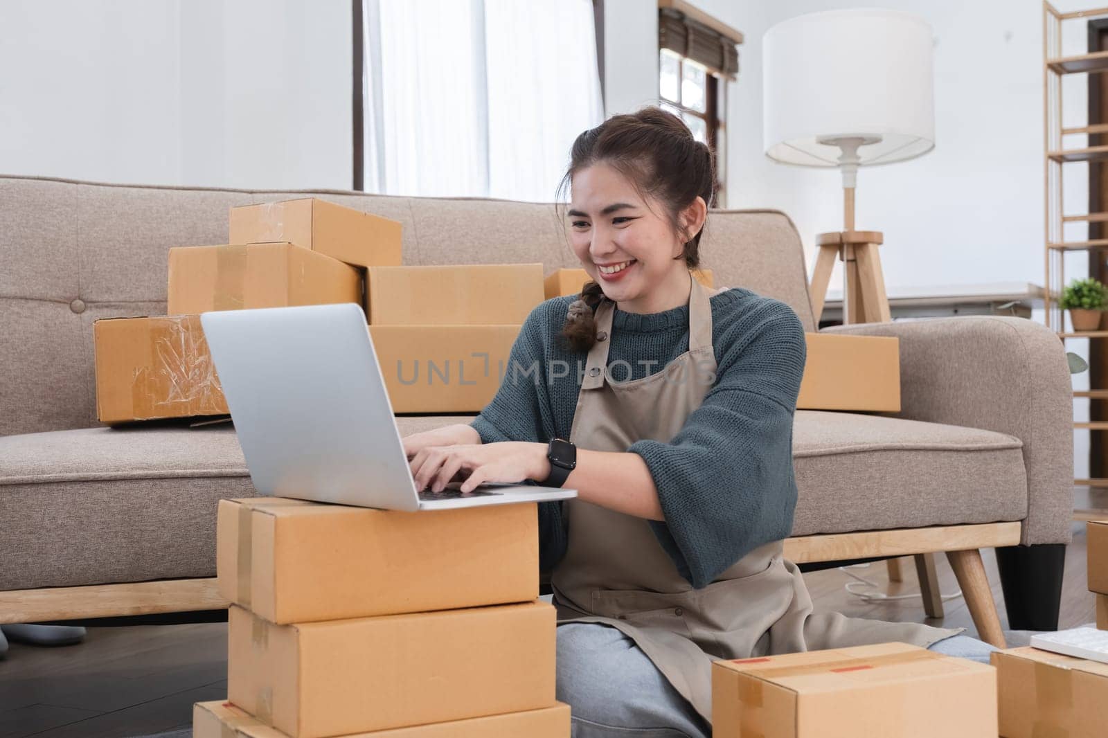 Young woman, small business owner selling products online, accepting online product orders via laptop, sits in a room with boxes of products. by wichayada