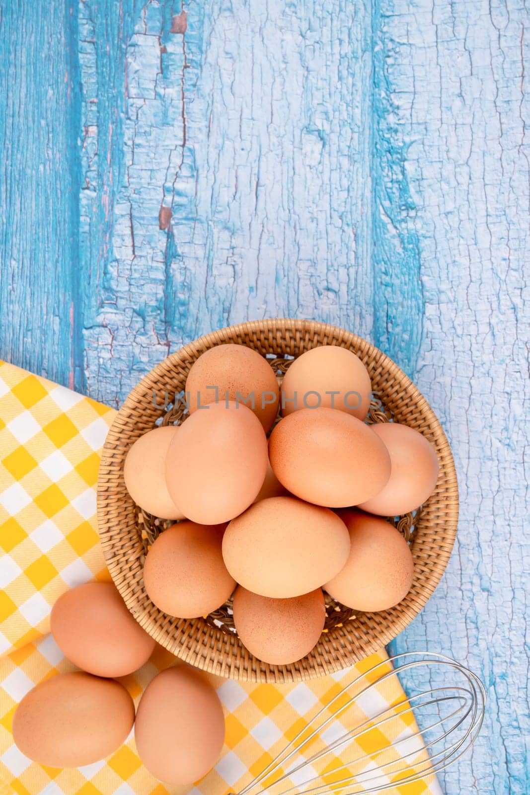 Overhead view of brown chicken eggs in weave basket and a whisk on blue wooden background.