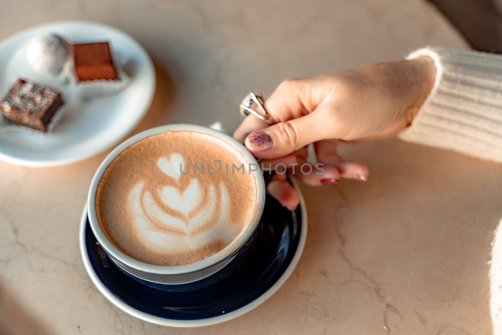 A cup of fresh cappuccino coffee in the hands of a woman on a fashionable background of a white marble table, next to a plate with sweets. Coffee addiction. Top view, flat lay. by Matiunina