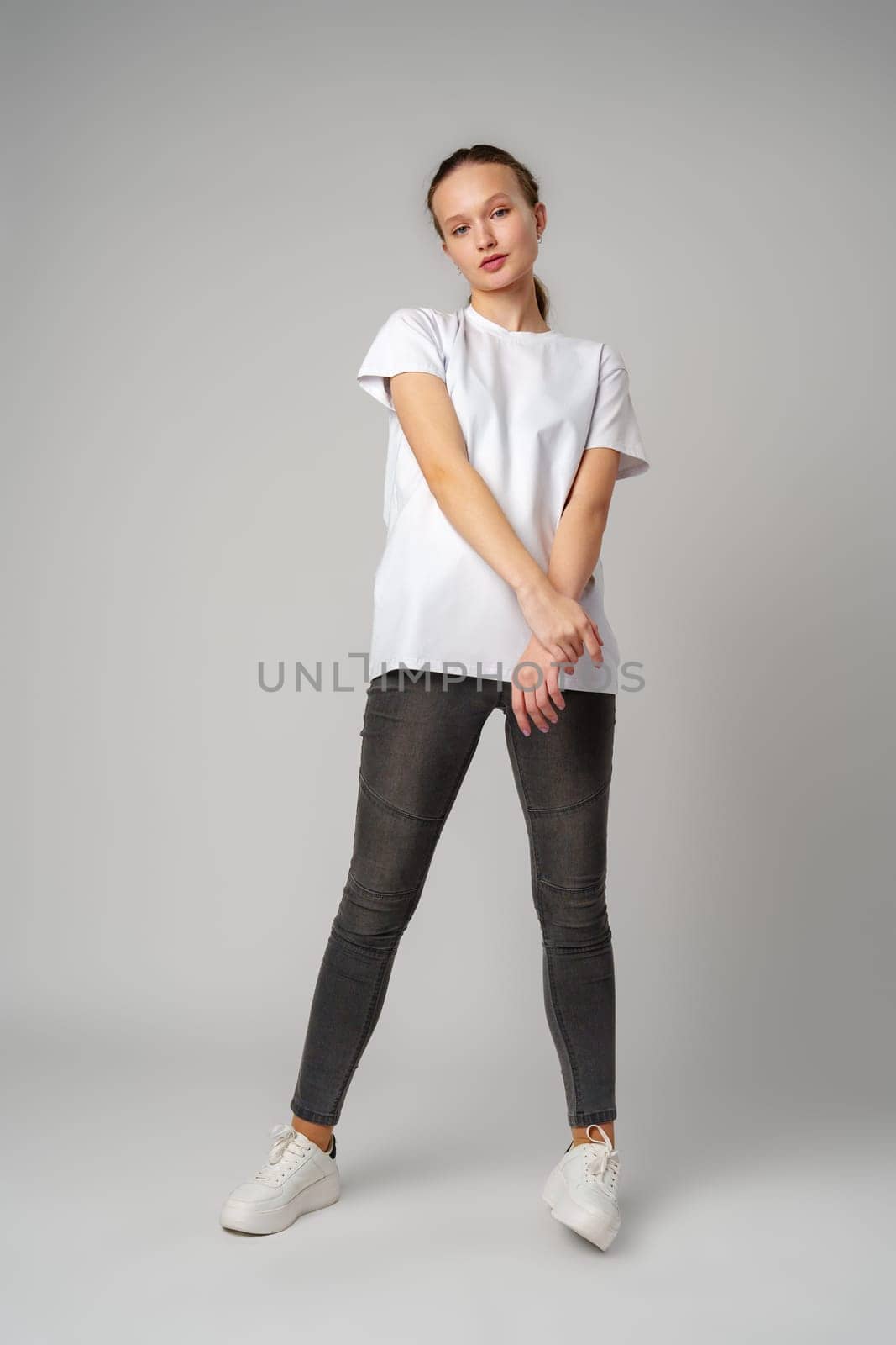 Beautiful young girl posing in white T-shirt and jeans on gray background by Fabrikasimf