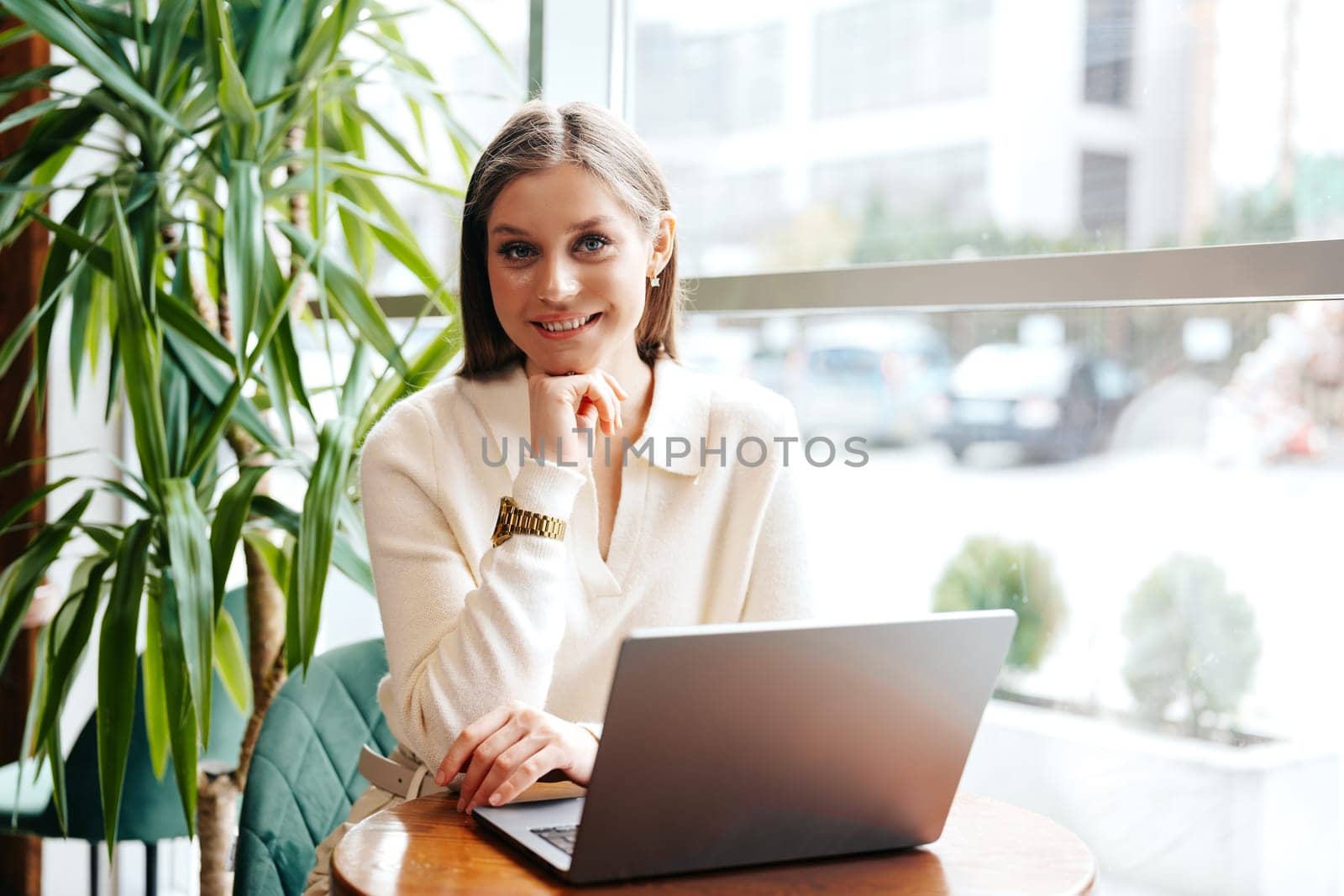 A cheerful young woman is seated at a wooden table in a well-lit cafe, working on her laptop. Her attention is momentarily diverted away from the screen as she smiles towards someone or something, giving off a pleasant and relaxed vibe amidst the indoor greenery that adorns the environment.