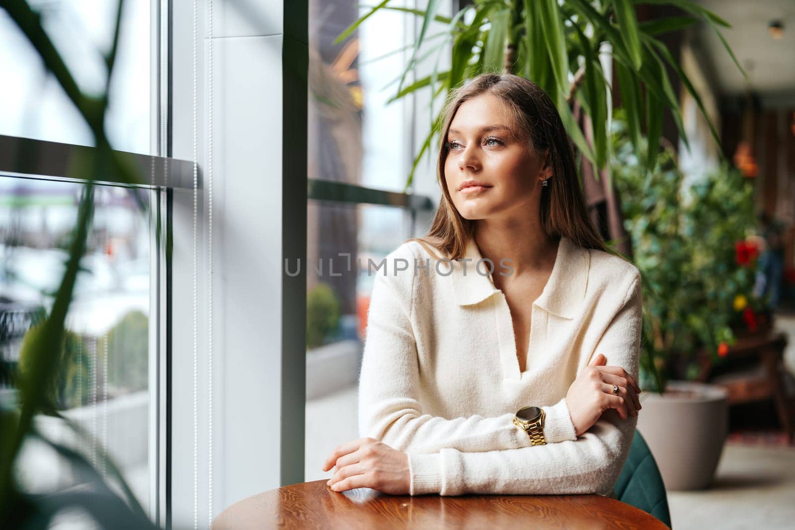 A woman is seated at a wooden table positioned in front of a large window. She appears to be focused on something outside, with natural light illuminating the scene.