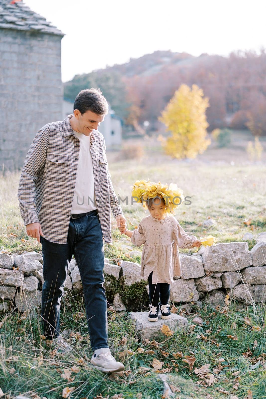 Little girl in a wreath of yellow leaves stands on a stone on the lawn of the house, holding her dad hand by Nadtochiy