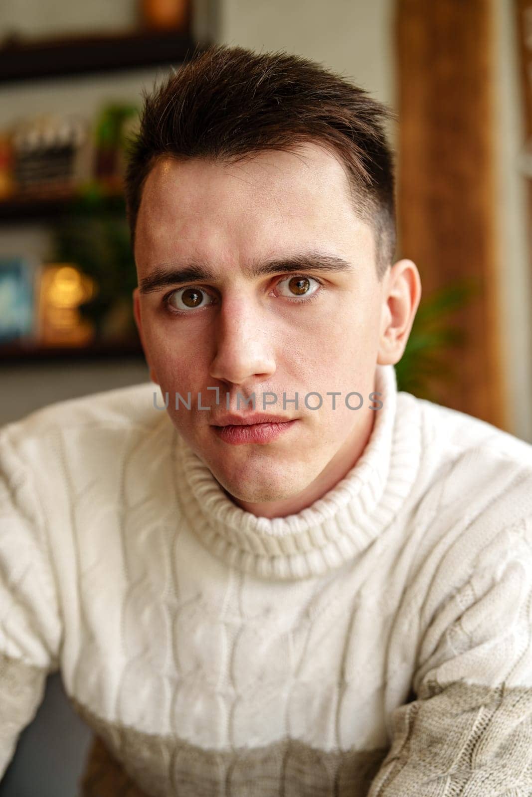 A male individual wearing a white sweater is directly facing the camera, making eye contact with the viewer. He appears to be posing for a portrait, with a neutral expression on his face.