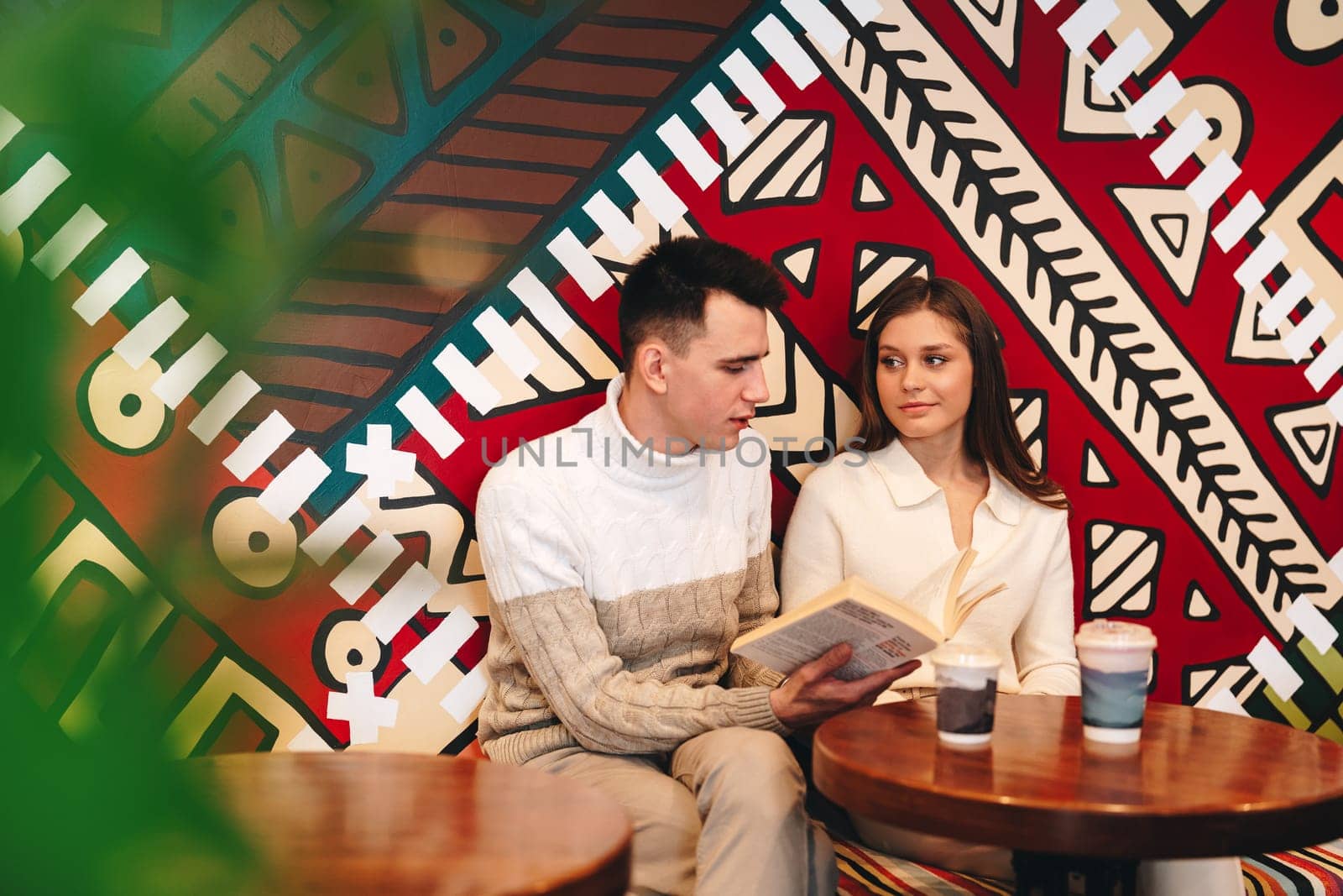 A man and a woman are seated at a wooden table, focused on reading a book. The man is wearing a blue shirt and glasses, while the woman has long hair and a pink sweater. The table is cluttered with books and a cup of coffee.