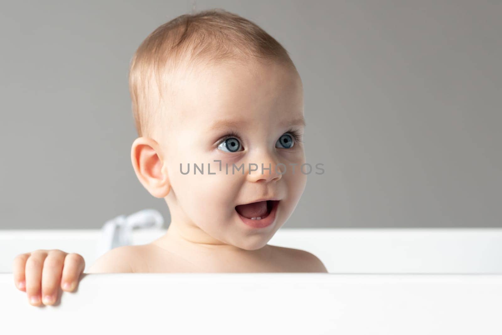 Close-up of the face of a baby laughing out loud and looking out of a crib with interest.