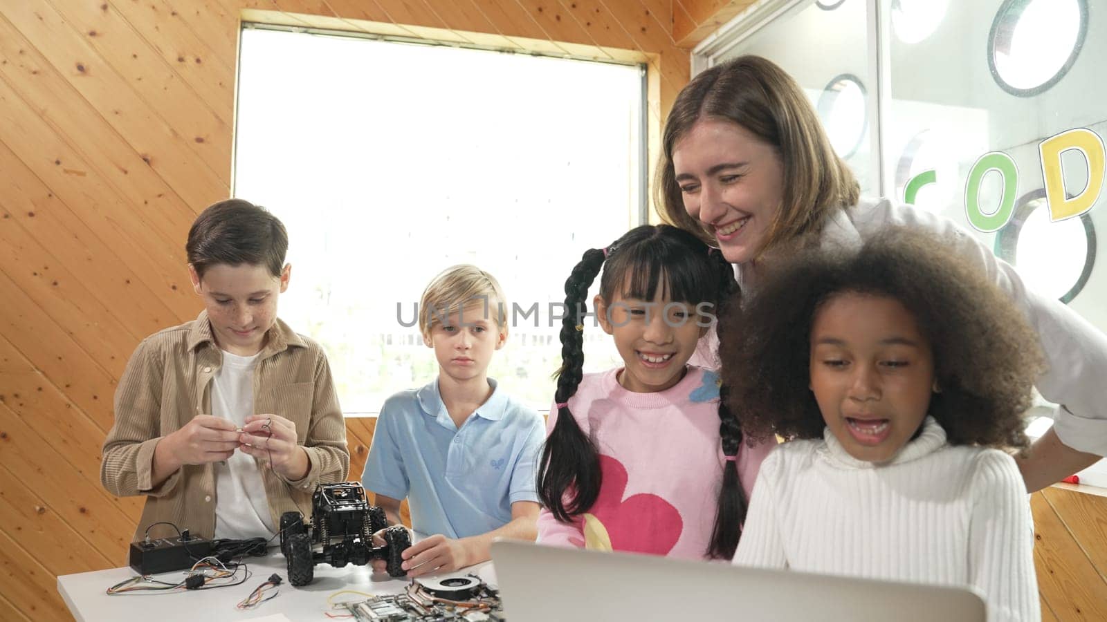 Caucasian teacher praise student while looking at learner work or presentation. Group of diverse student looking at presentation and fixing motherboard at table with chips and wires placed. Erudition.