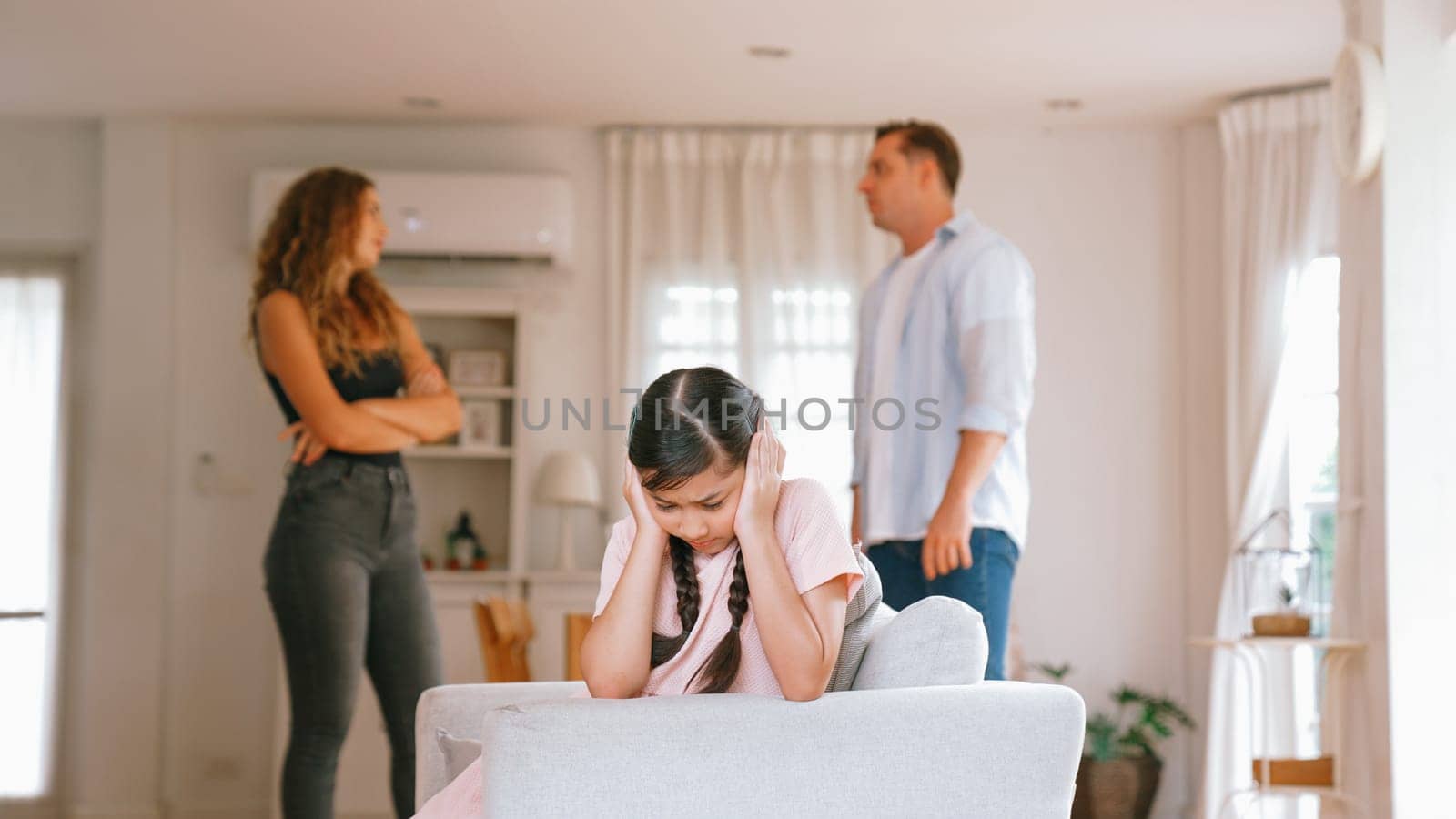 Annoyed and unhappy young girl sitting on sofa trapped in middle of tension by her parent argument in living room. Unhealthy domestic lifestyle and traumatic childhood develop to depression Synchronos
