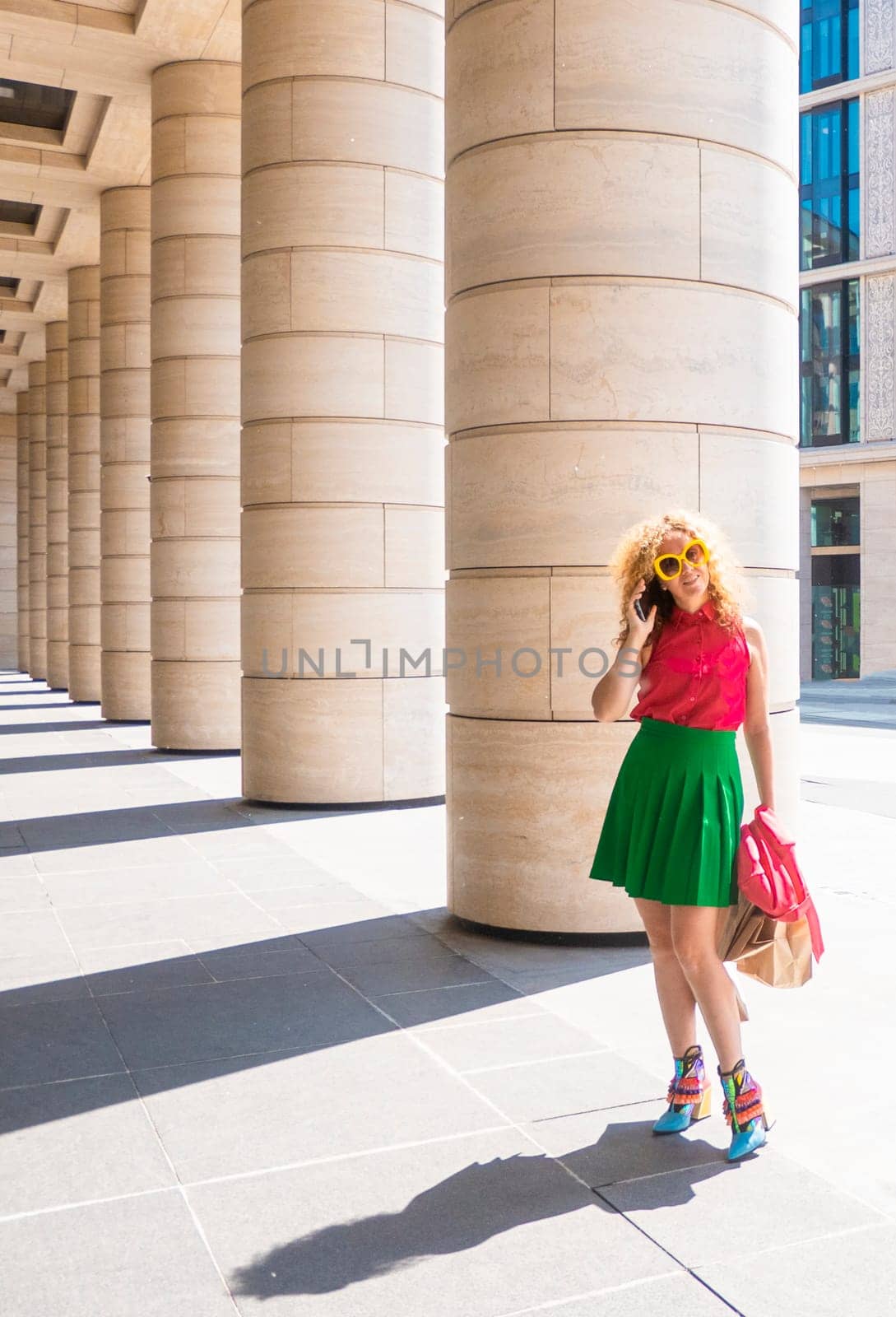 Content fashion female with bag walking on pavement near stone columns in sunlight and talk cell phone. by kajasja