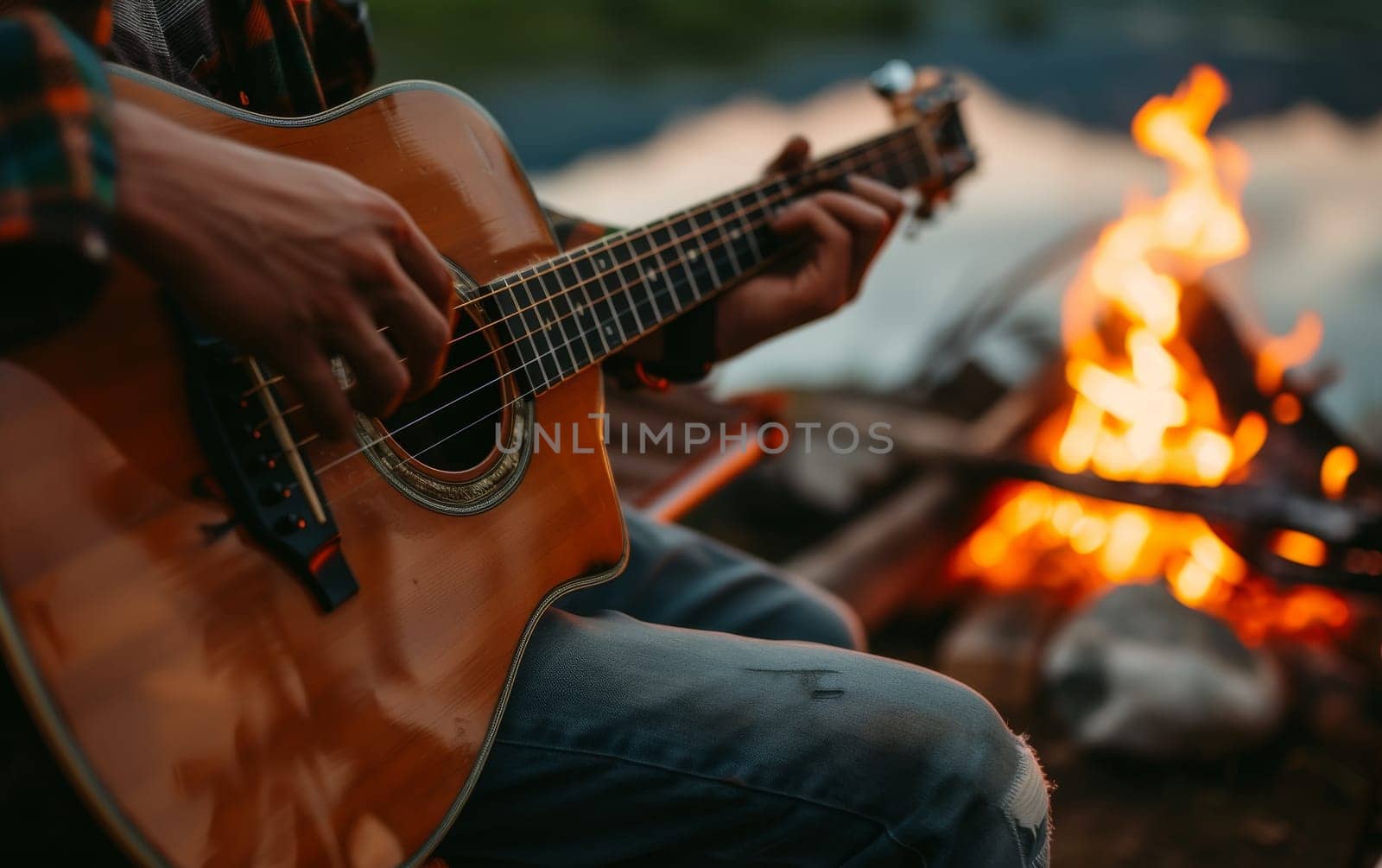Person playing an acoustic guitar by a campfire near a tranquil lake at dusk