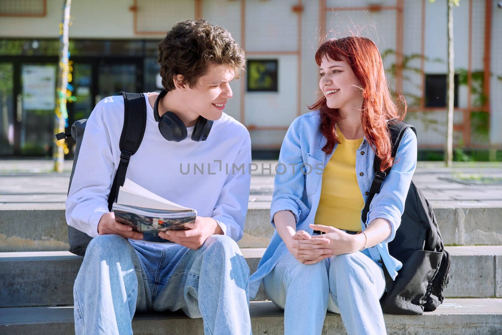 Teenage college students guy and girl talking, sitting outdoor near educational building. by VH-studio