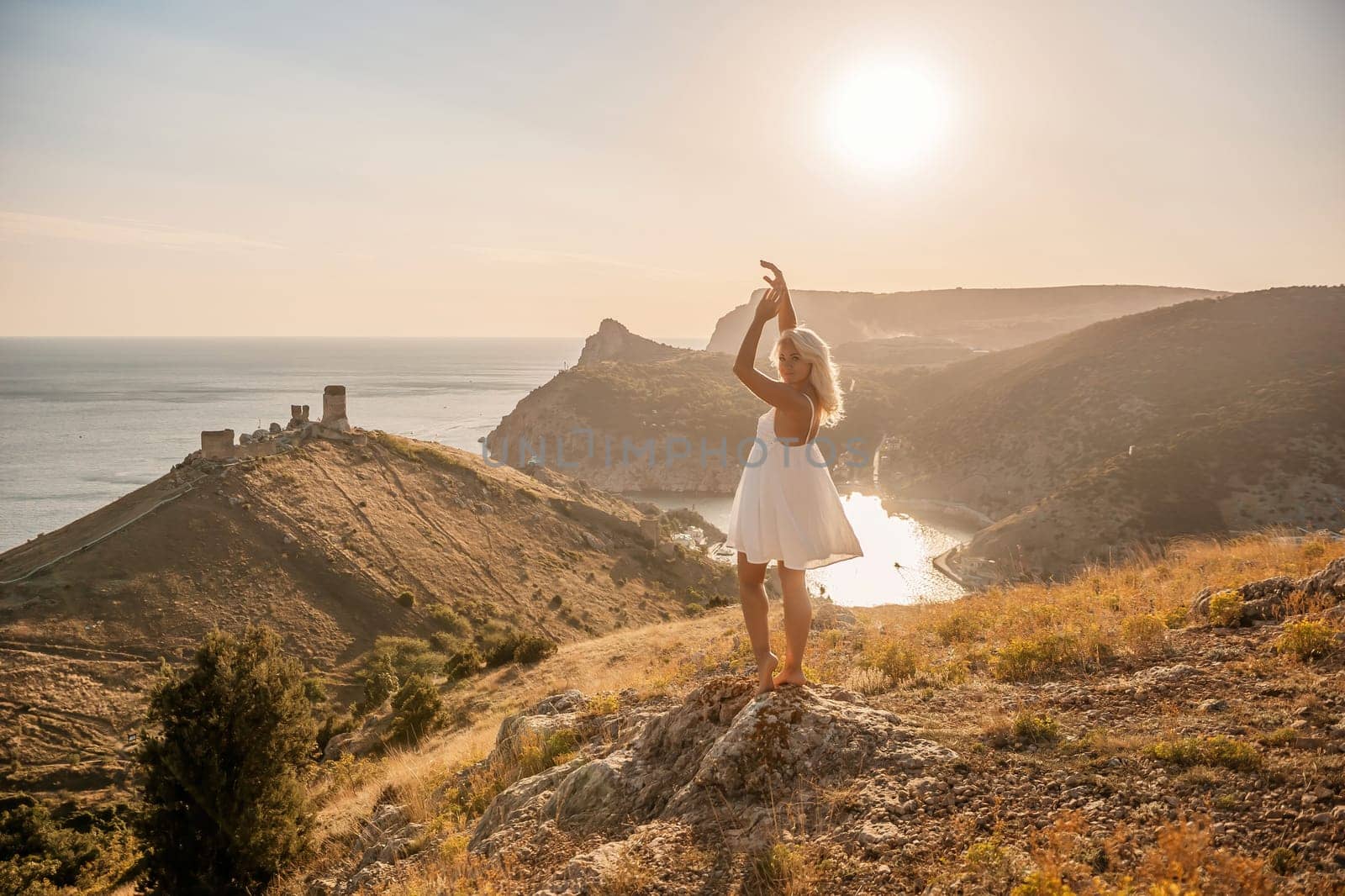 A woman stands on a hill overlooking a body of water. She is wearing a white dress and she is enjoying the view