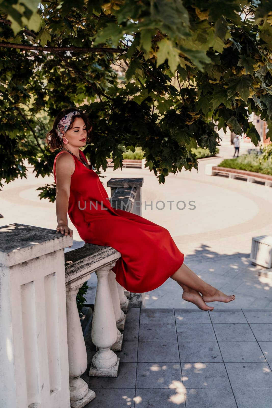 A pretty woman in a red silk dress and a bandage on her head smiles against the background of the leaves of a tree. She is leaning on the coop and looking into the camera. Vertical photo