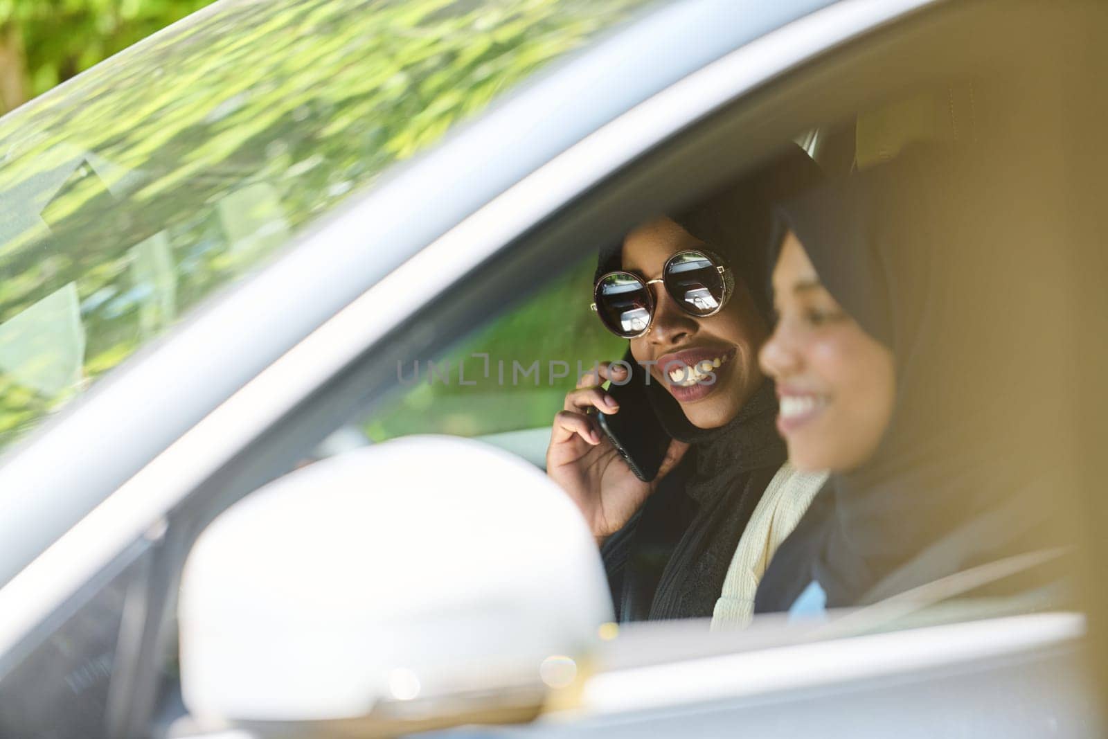 Two Muslim women wearing hijab converse on a smartphone while traveling together in a car through the by dotshock