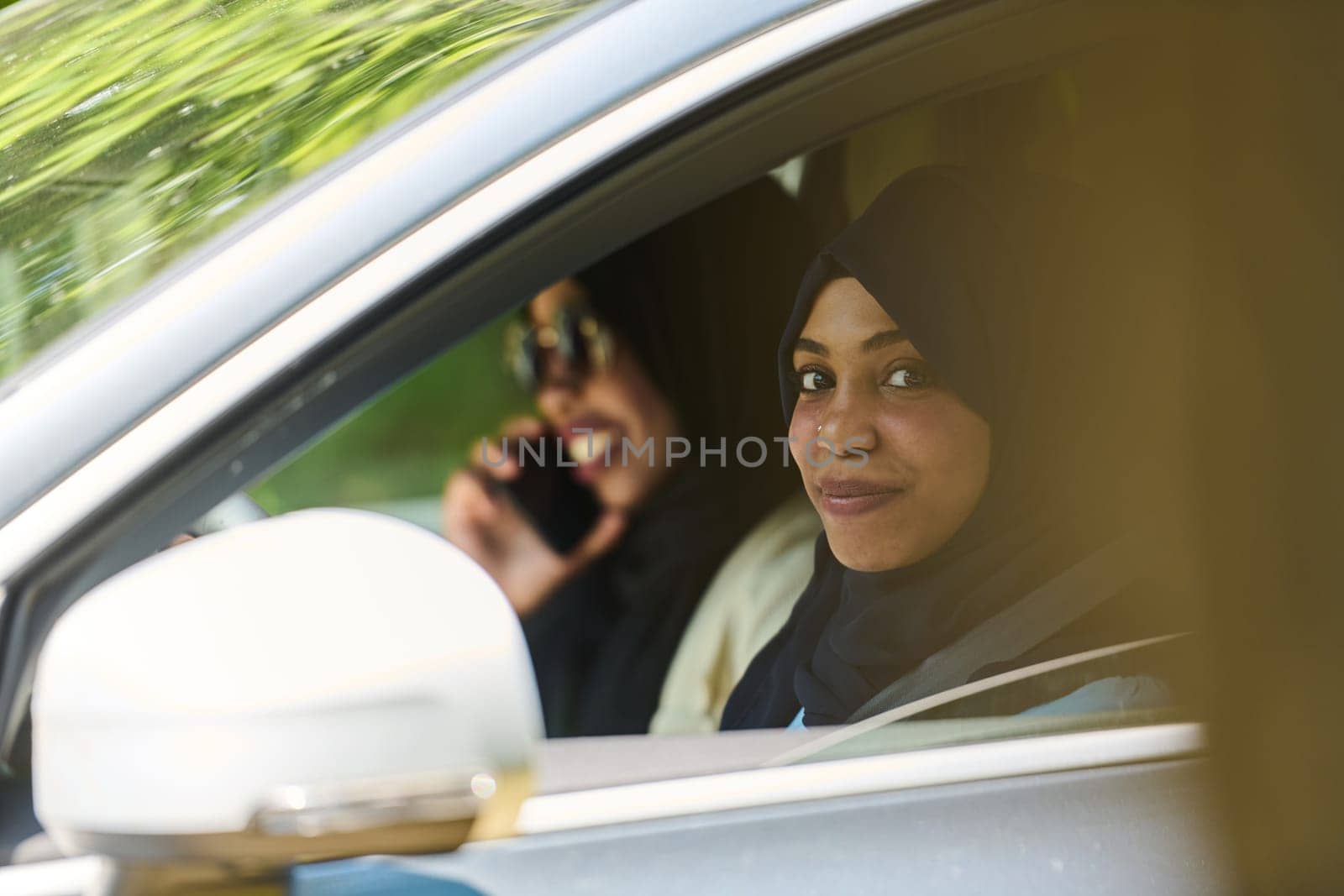 Two Muslim women wearing hijab converse on a smartphone while traveling together in a car through the.
