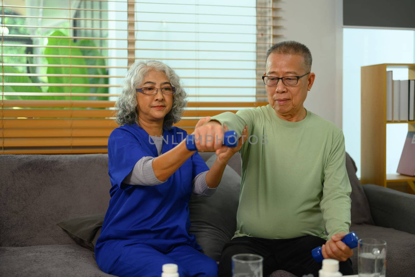 Professional physiotherapist helping elderly patient doing exercises with light weight dumbbell. by prathanchorruangsak