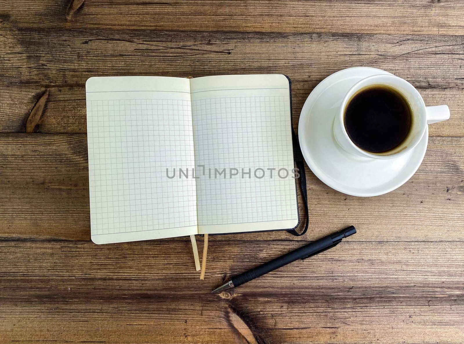 Coffee in a cup and a notebook with a pen on the table, top view. Coffee in a cup with a saucer and a notebook with a pen on a wooden table.